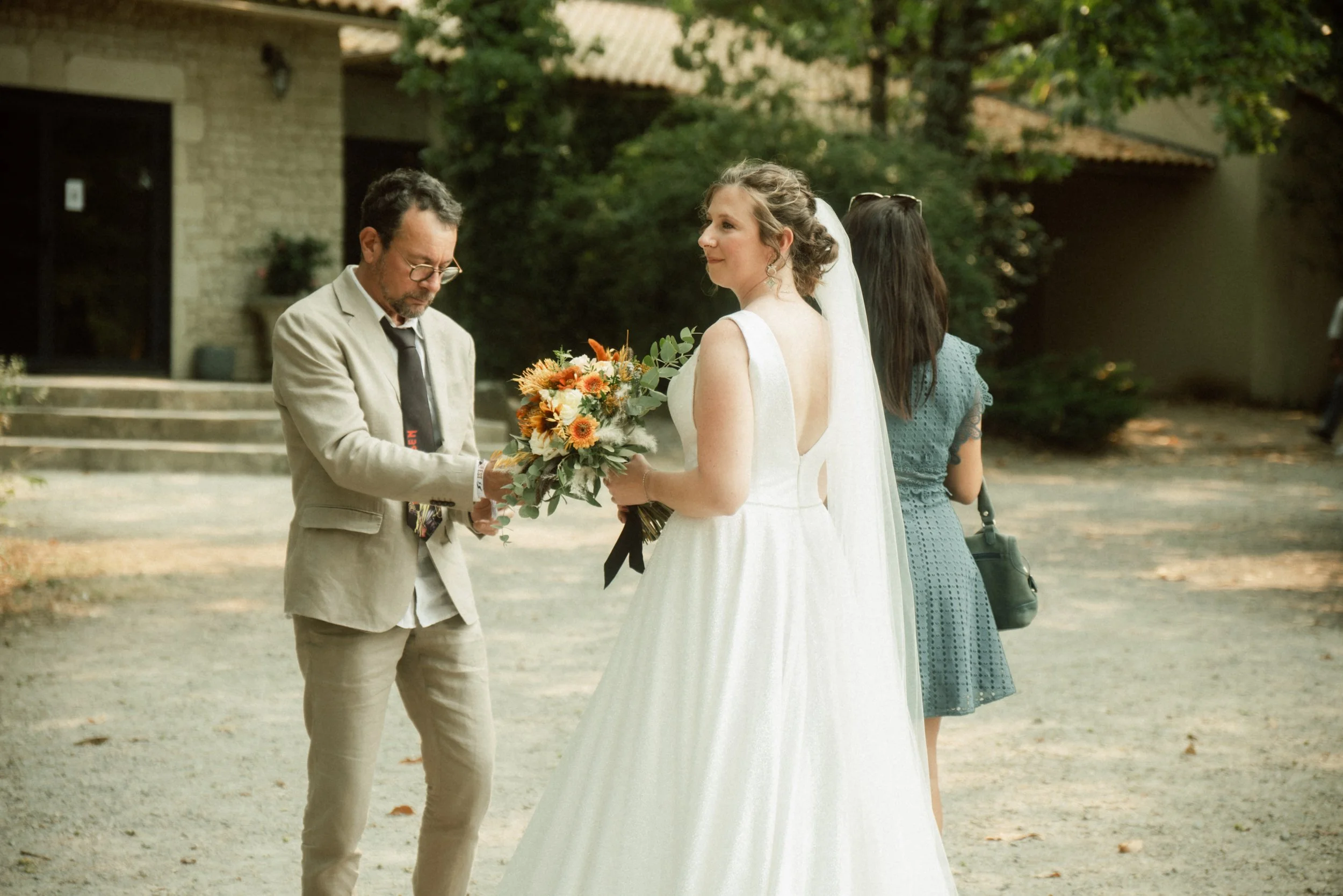 Une mariée en robe blanche reçoit un bouquet de fleurs cultivées, tandis qu'un homme en costume beige lui tend le bouquet, dans un cadre extérieur avec des arbres et une maison en arrière-plan.