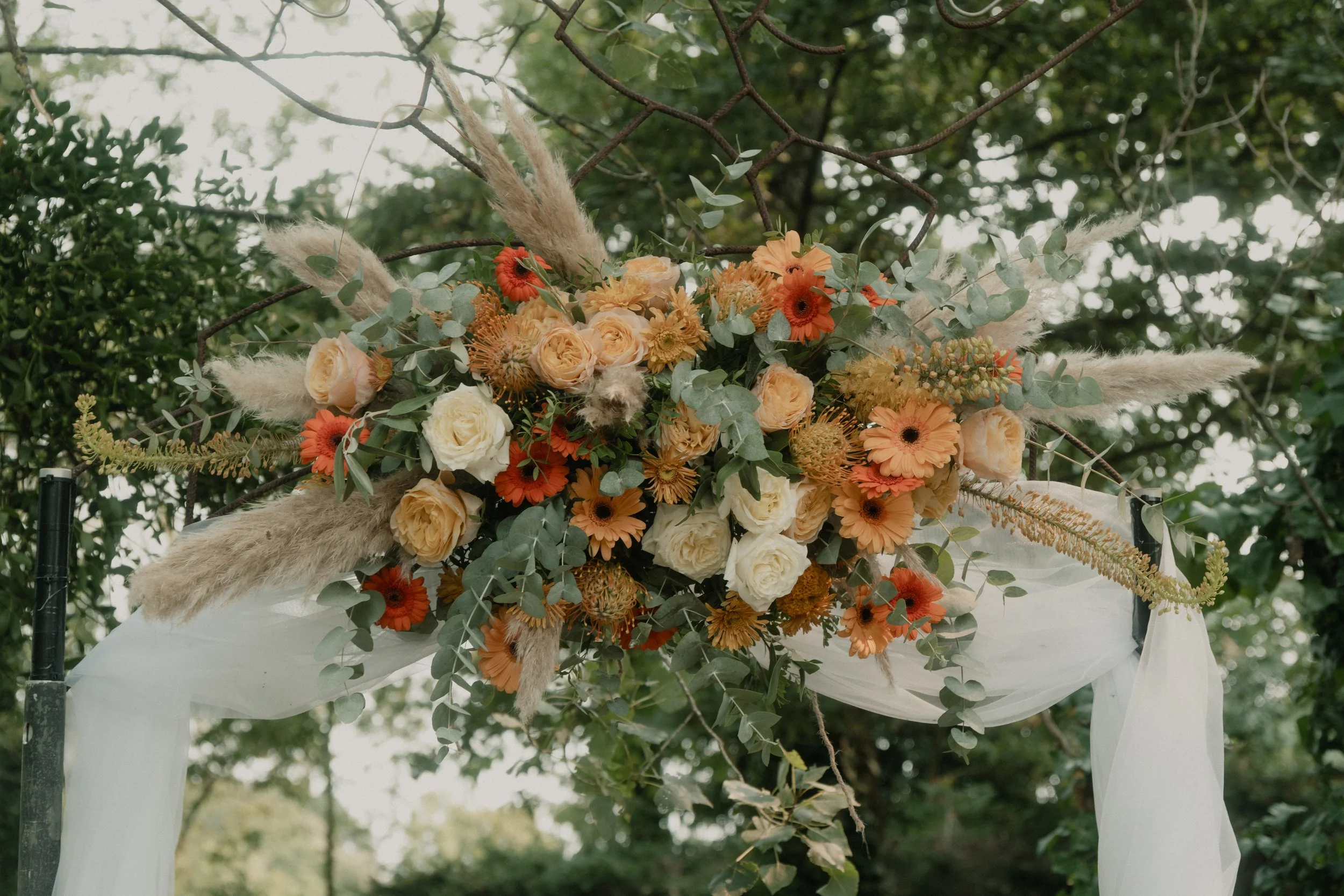 Arrangement floral avec roses blancs et jaunes, gerberas orange, eucalyptus et pampas, décorant un arc de mariage en extérieur.