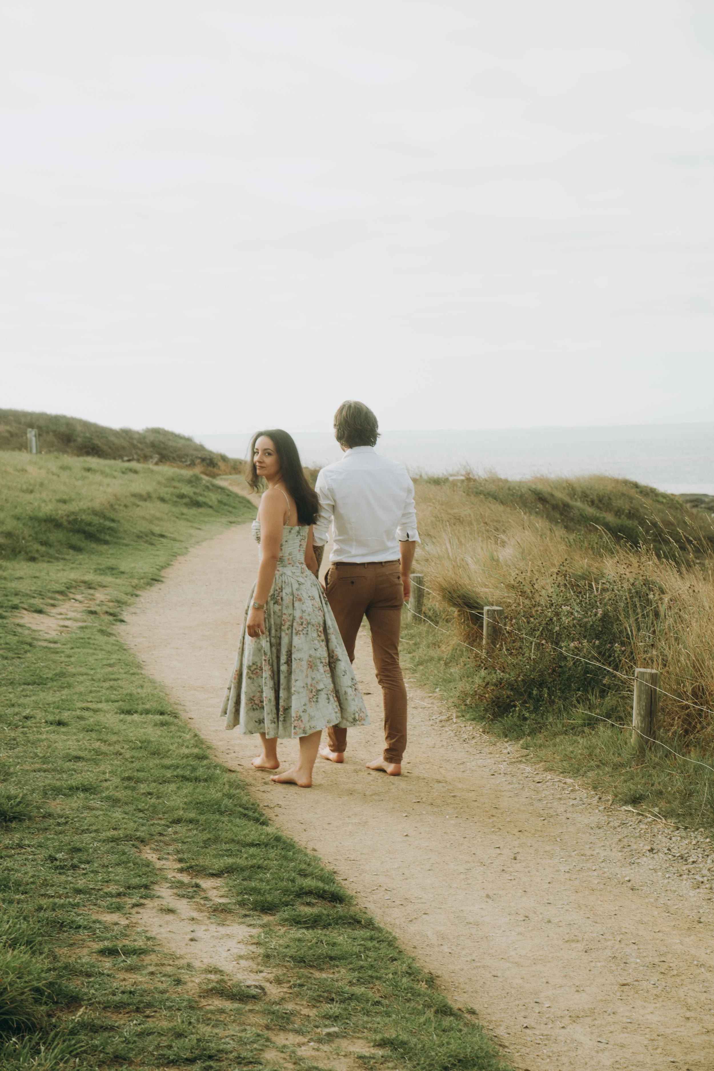 Un couple marche pieds nus le long d'un chemin côtier. La femme porte une robe fleurie longue et regarde en arrière. L'homme porte une chemise blanche et un pantalon marron. La scène se déroule sous un ciel nuageux, entourée d'herbes hautes et de la 