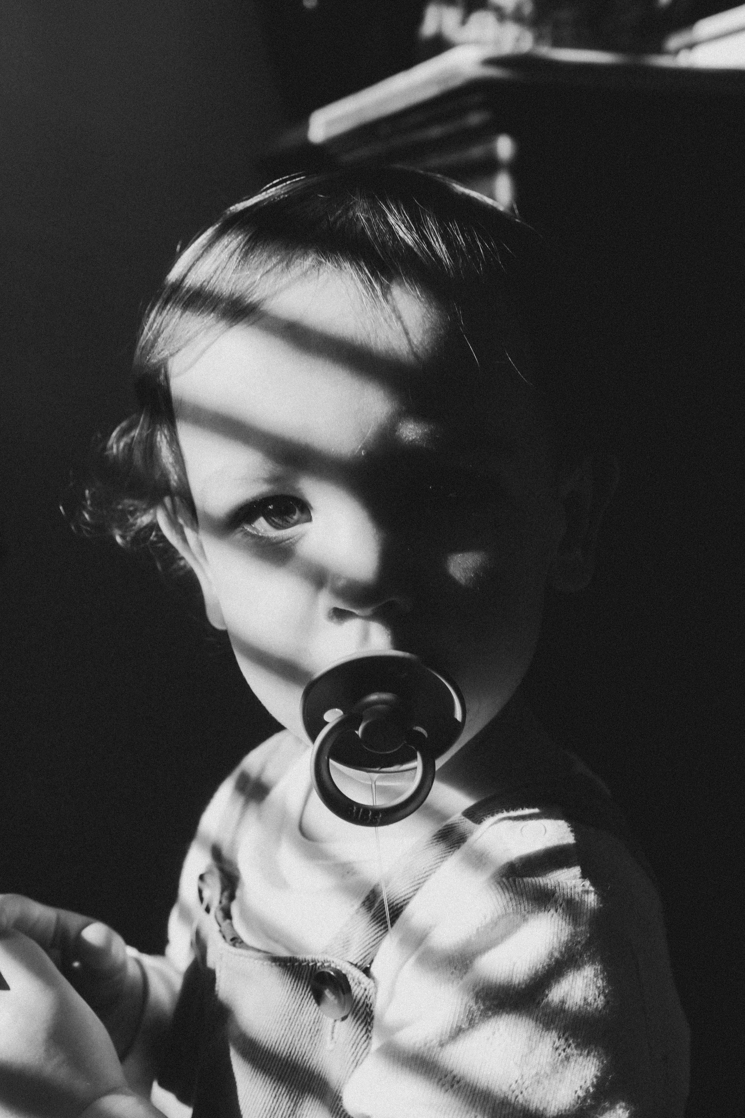 Un jeune enfant avec un tuteur en cheveux courts et un piercing à la lèvre, portant un vêtement à manches longues, regarde vers la caméra. La photo est en noir et blanc avec des lignes d'ombre sur le visage.