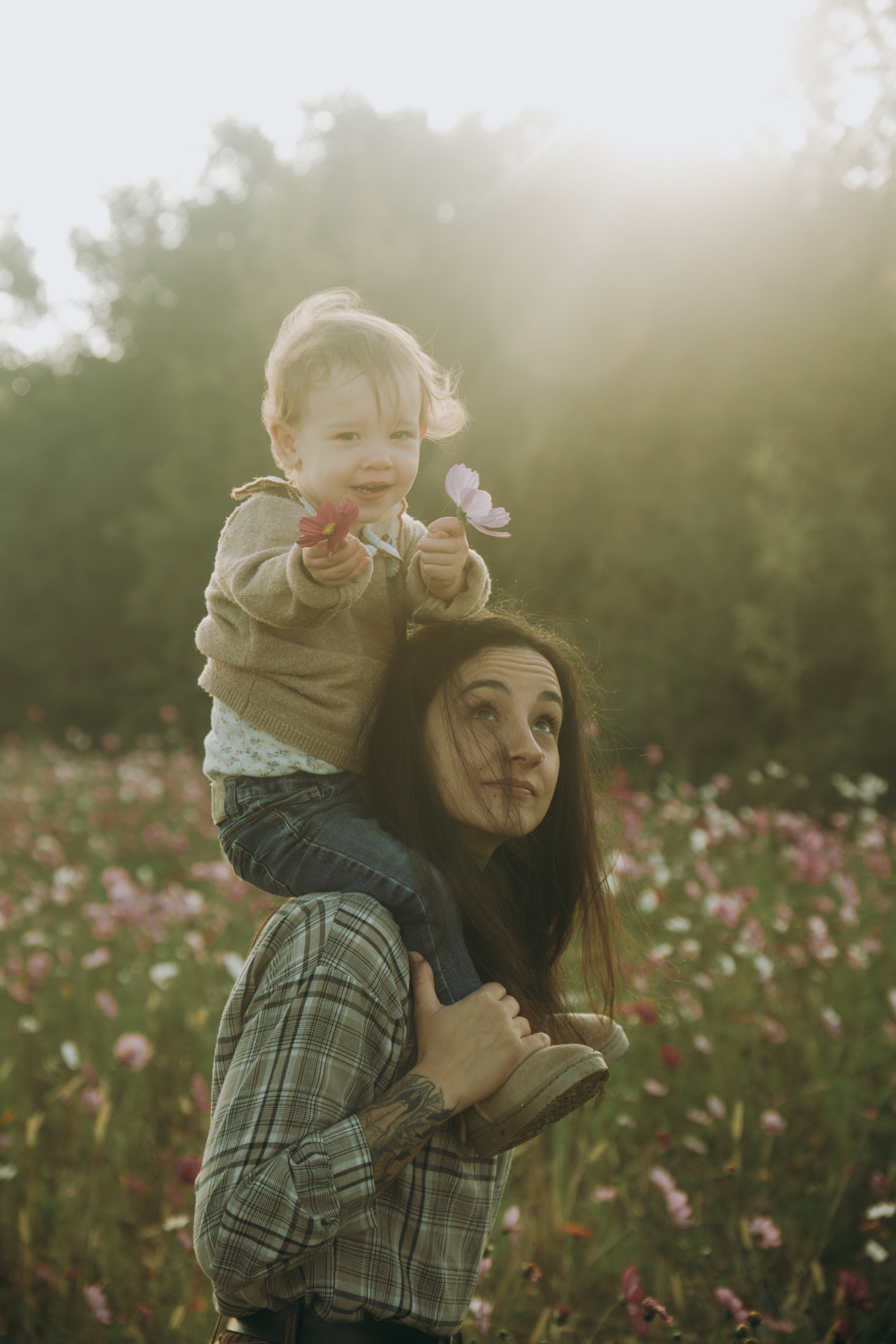 Une femme portant un enfant sur ses épaules dans un champ de fleurs, tenant des fleurs dans ses mains, avec des arbres au fond, sous une lumière de soleil douce.