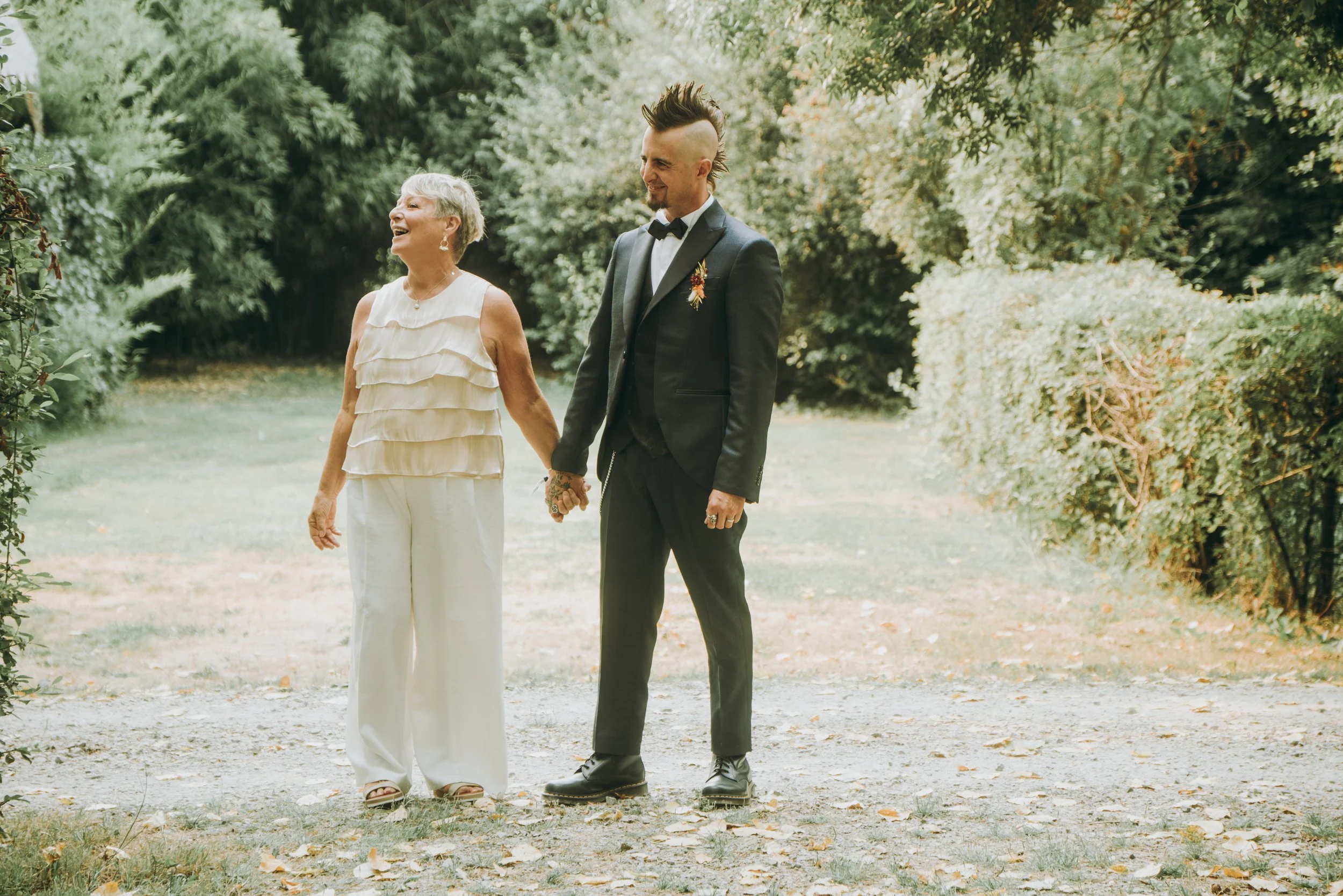 Une femme âgée et un homme jeune, vêtus élégamment, se tiennent dans un parc, tenant la main et souriant dans un cadre naturel avec des arbres.
