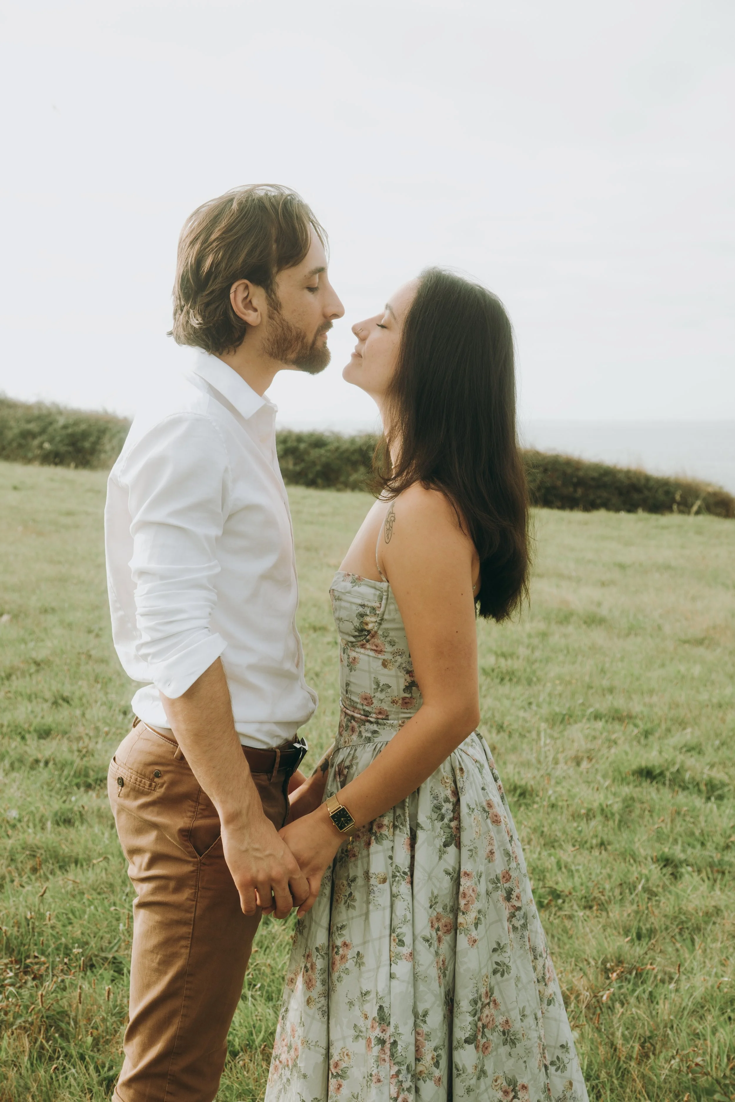 Un couple se tenant par la main et se regardant tendrement dans un champ vert avec l'océan en arrière-plan.