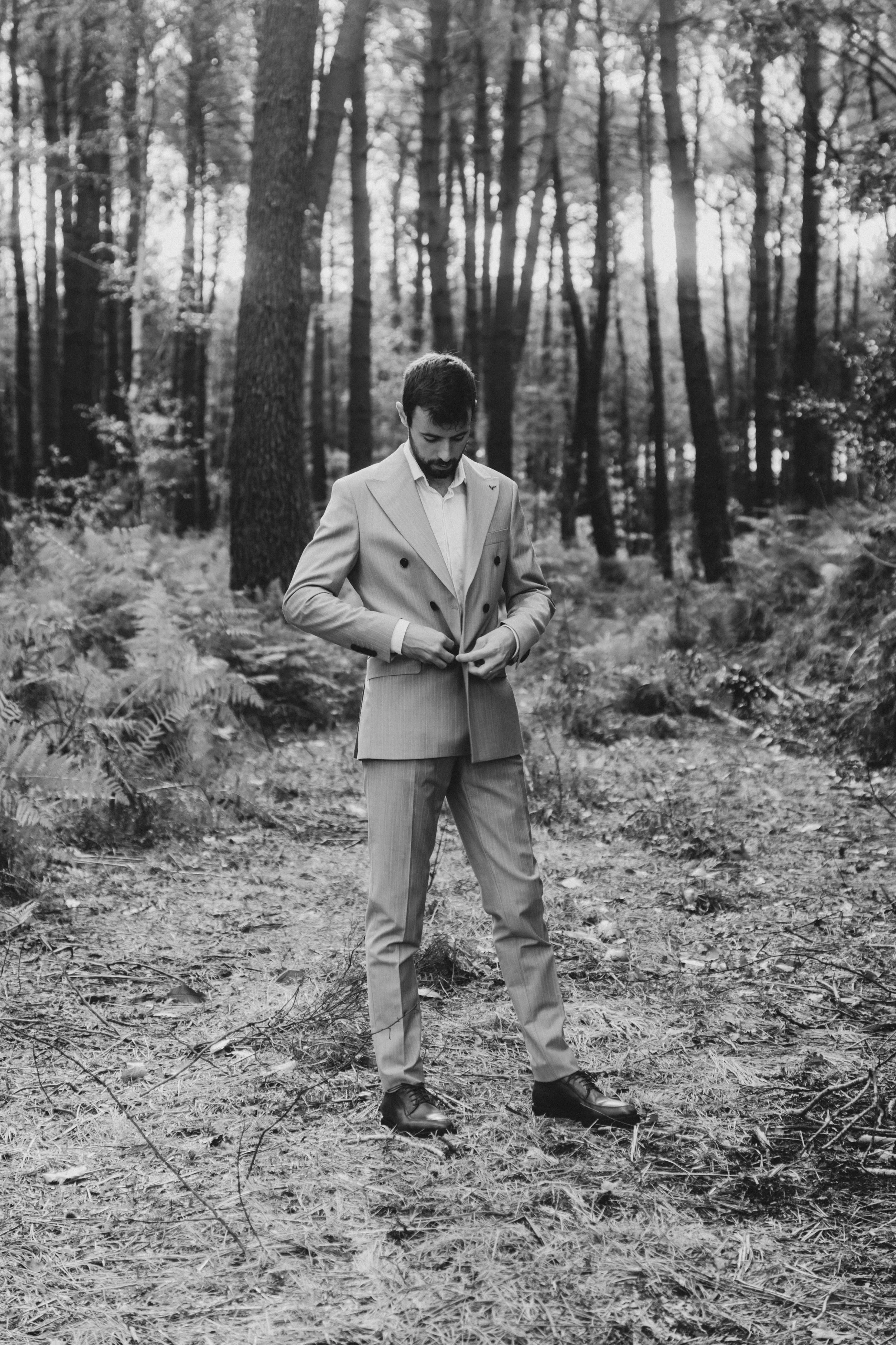 Un homme en costume dans une forêt, en train de boutonner sa veste.