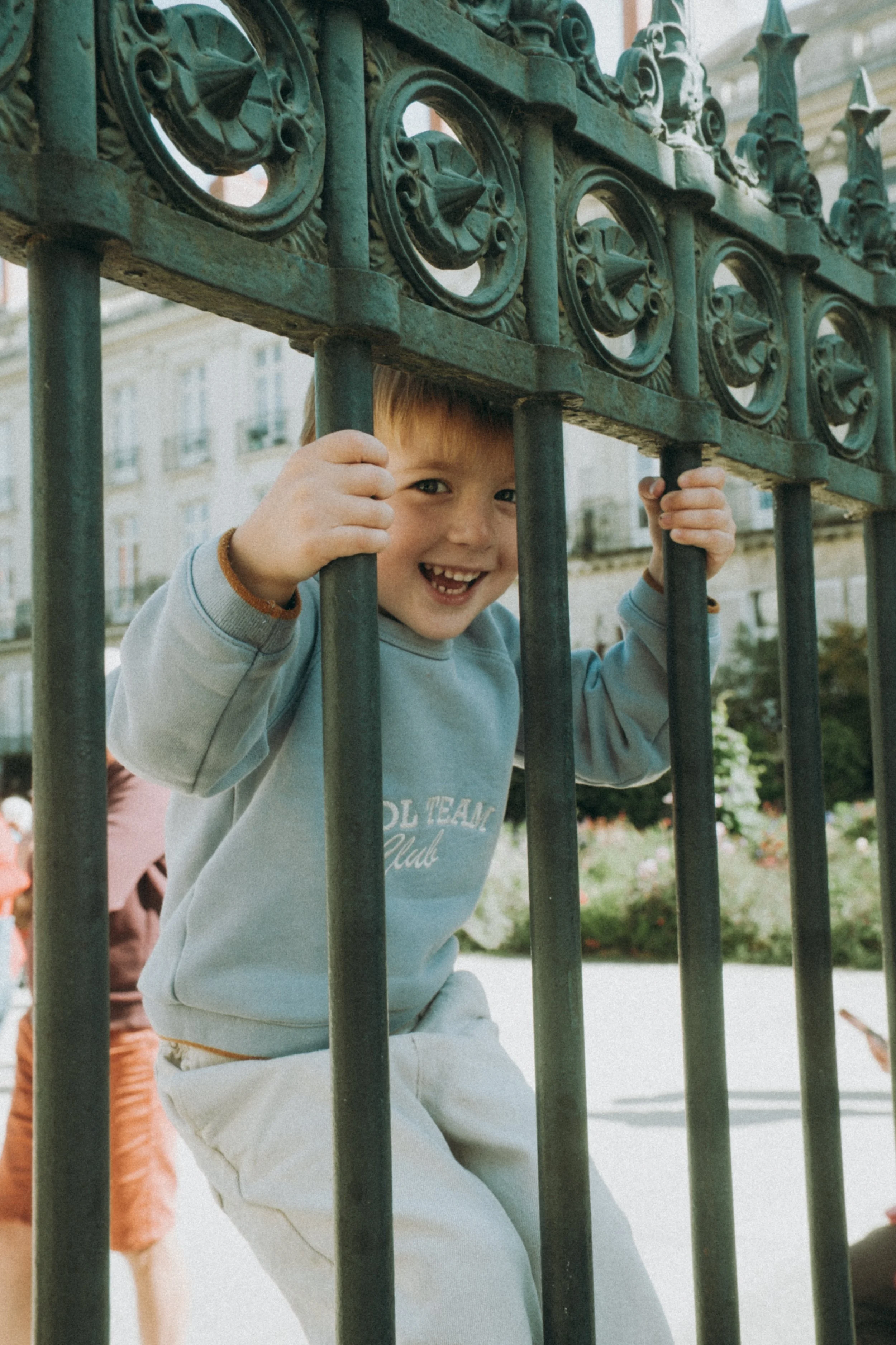 Un enfant souriant joue derrière une barrière en fer forgé dans un parc urbain, avec un bâtiment résidentiel en arrière-plan.