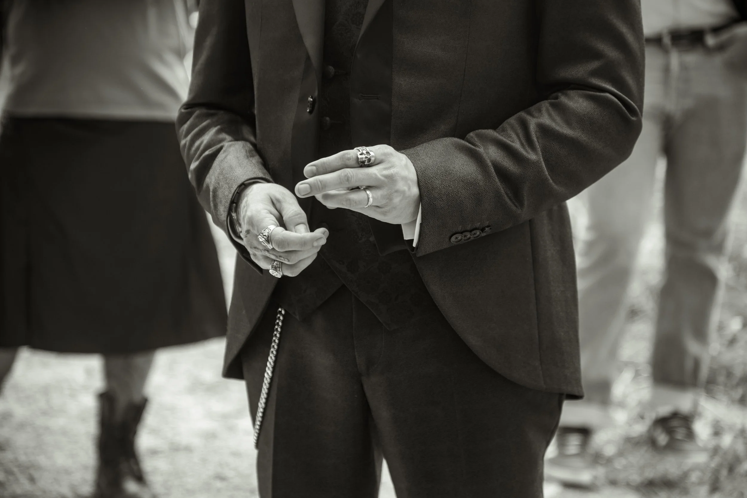 Une personne en costume sombre ajustant ses doigts, portant plusieurs bagues, avec d'autres personnes floues à l'arrière-plan.