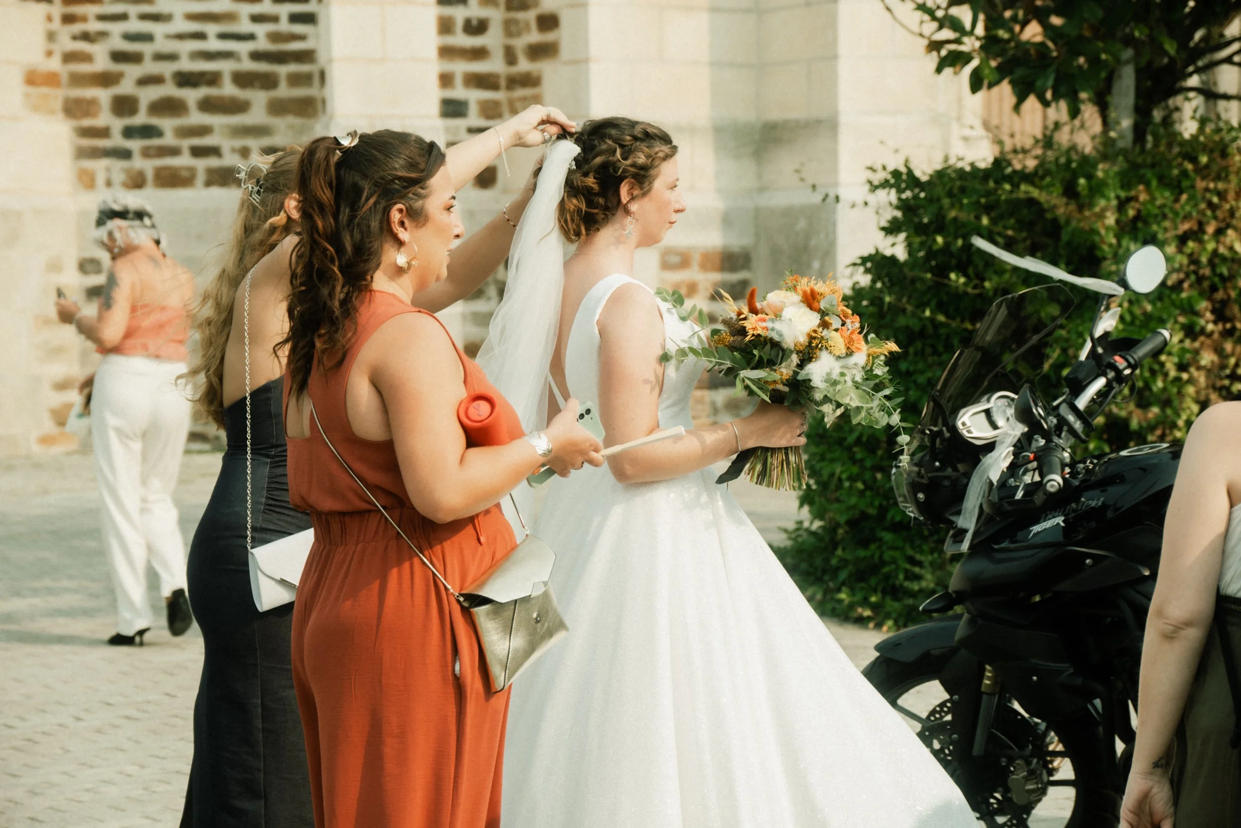 Une mariée habillée en robe blanche tient un bouquet de fleurs, entourée de femmes qui l'aident à préparer ses cheveux, dans un cadre extérieur avec un bâtiment en pierre en arrière-plan.