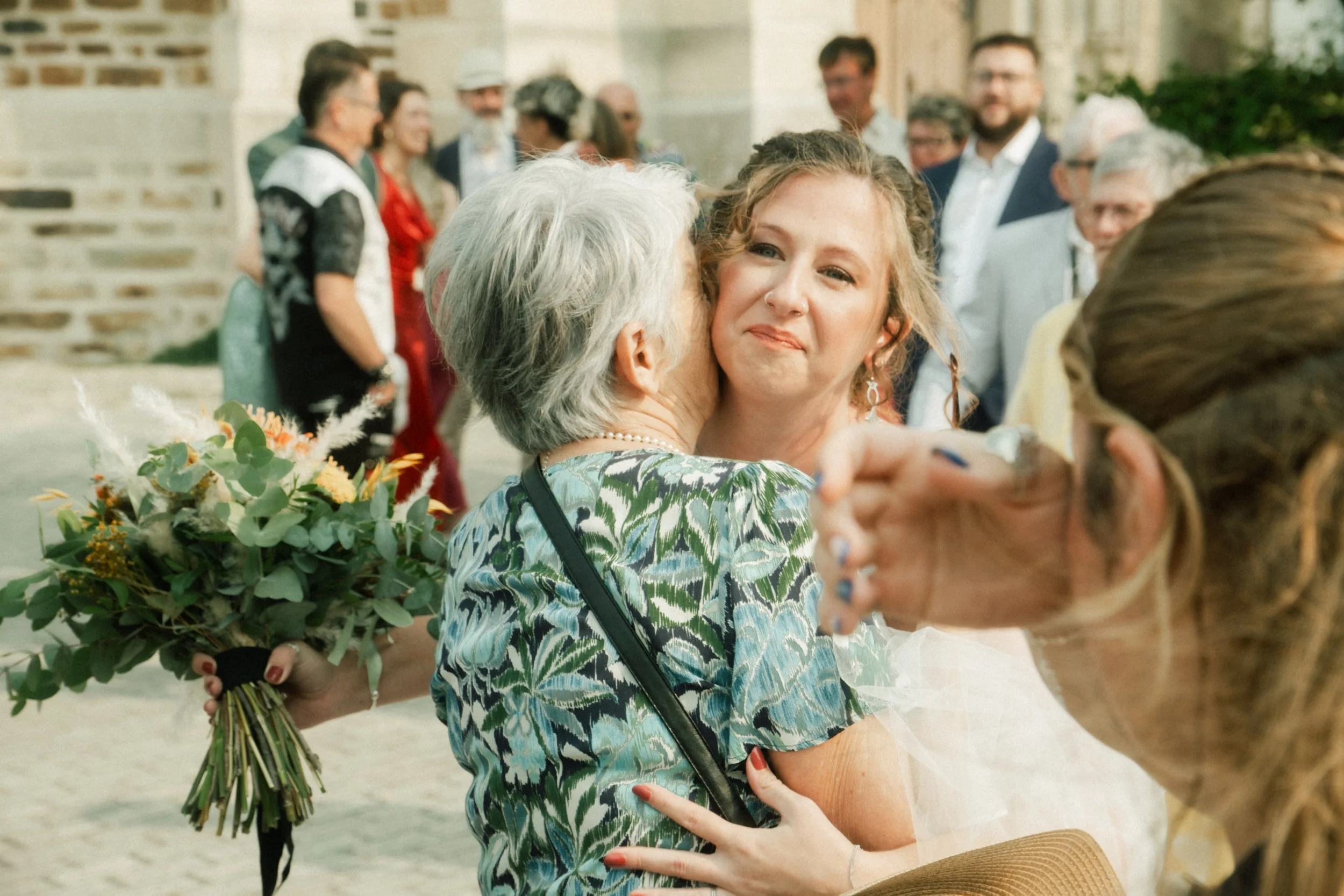 Une jeune femme heureuse embrassant une femme plus âgée, lors d'une célébration en plein air avec plusieurs personnes en arrière-plan.