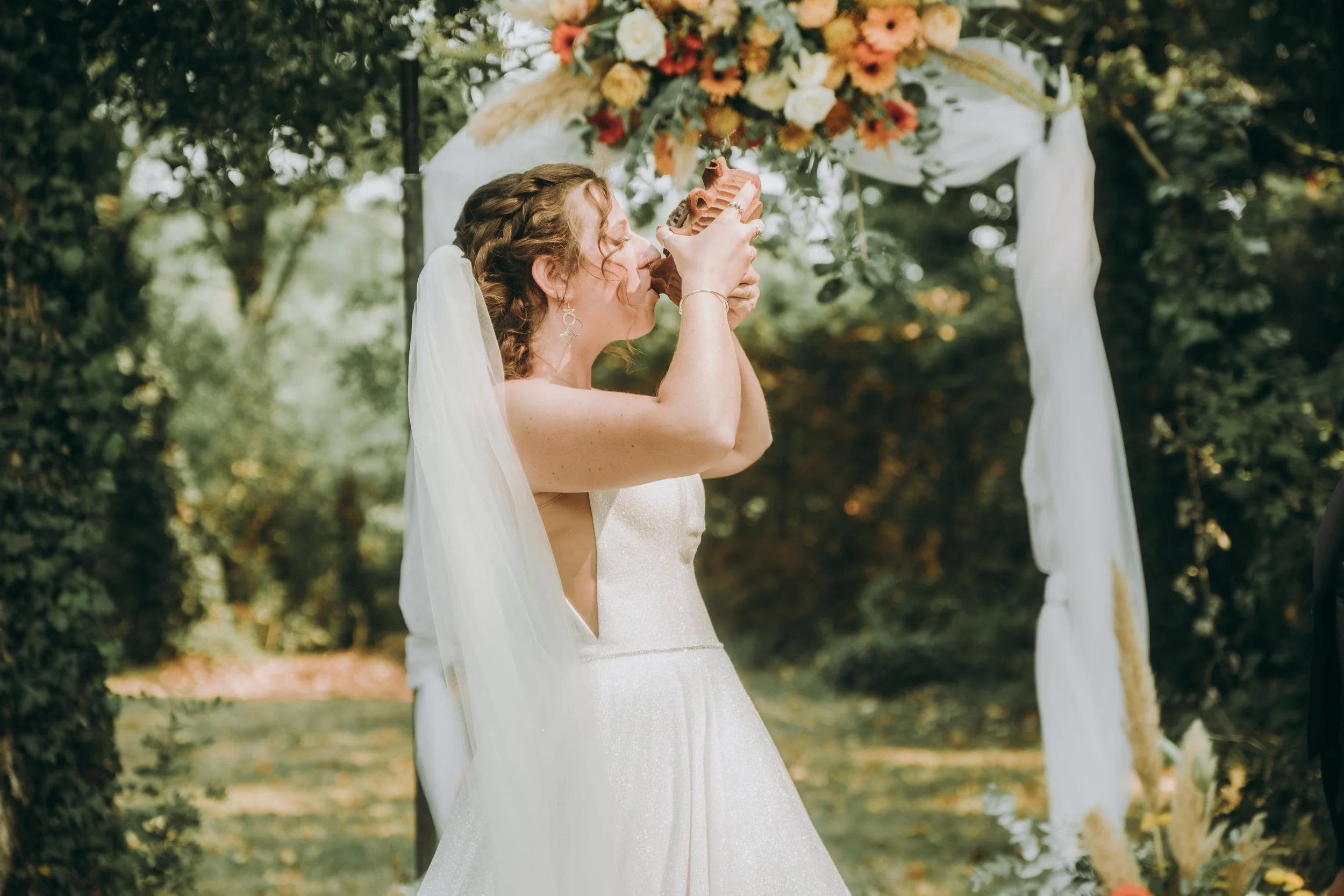 Une femme en robe de mariage avec un voile, célébrant un mariage, tenant un objet en forme de silhouette, sous une arche décorée de fleurs dans un environnement extérieur.