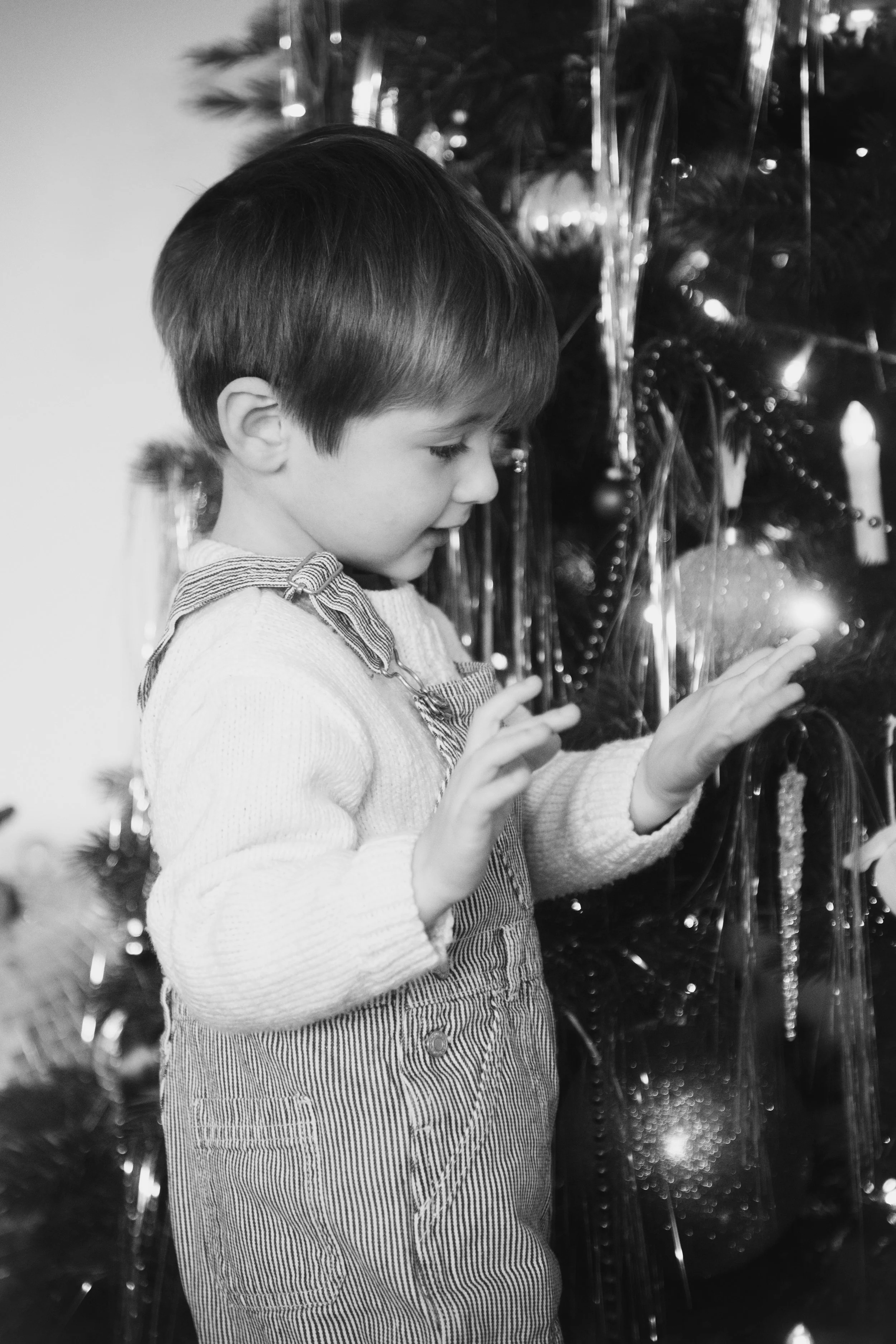 Un jeune garçon regarde et touche un arbre de Noël décoré avec des boules et guirlandes, en noir et blanc.