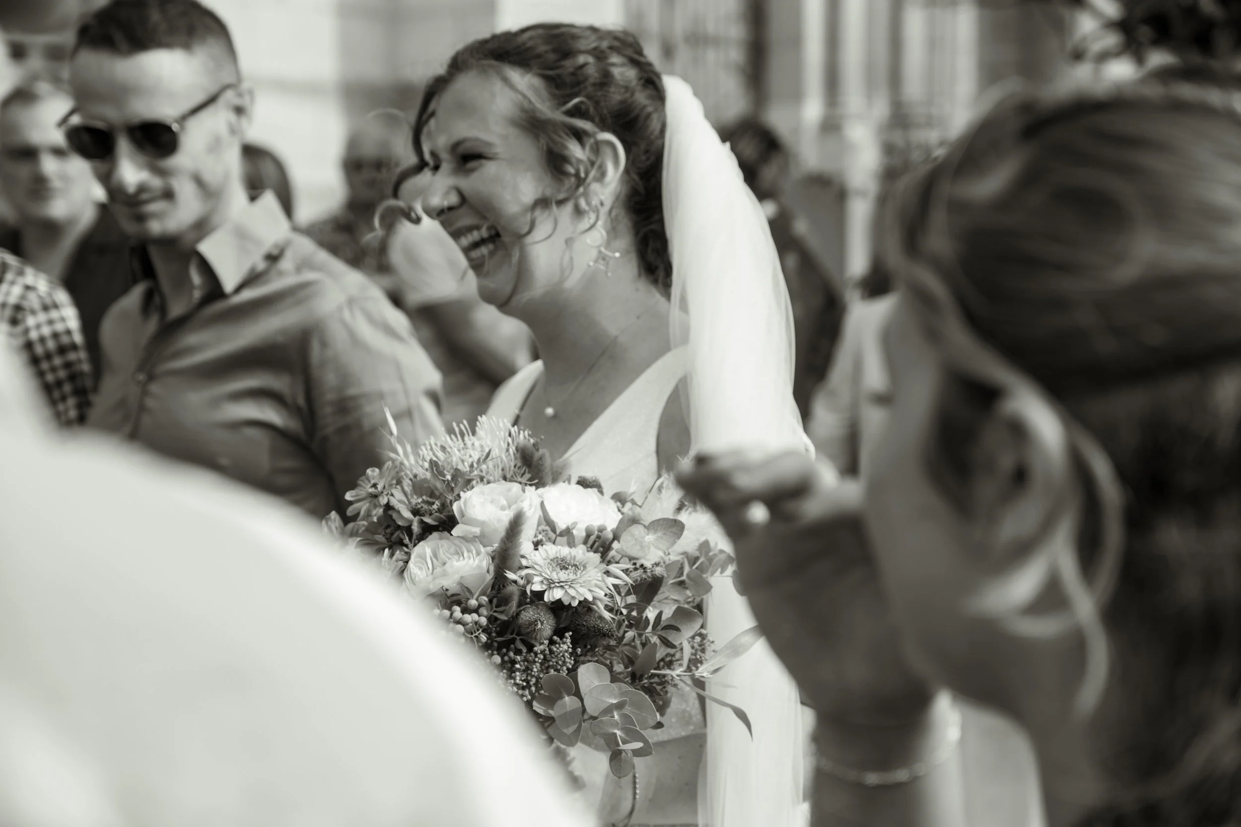 Une femme en robe de mariage sourit en tenant un bouquet de fleurs lors d'une célébration, entourée de plusieurs personnes. L'image est en noir et blanc.