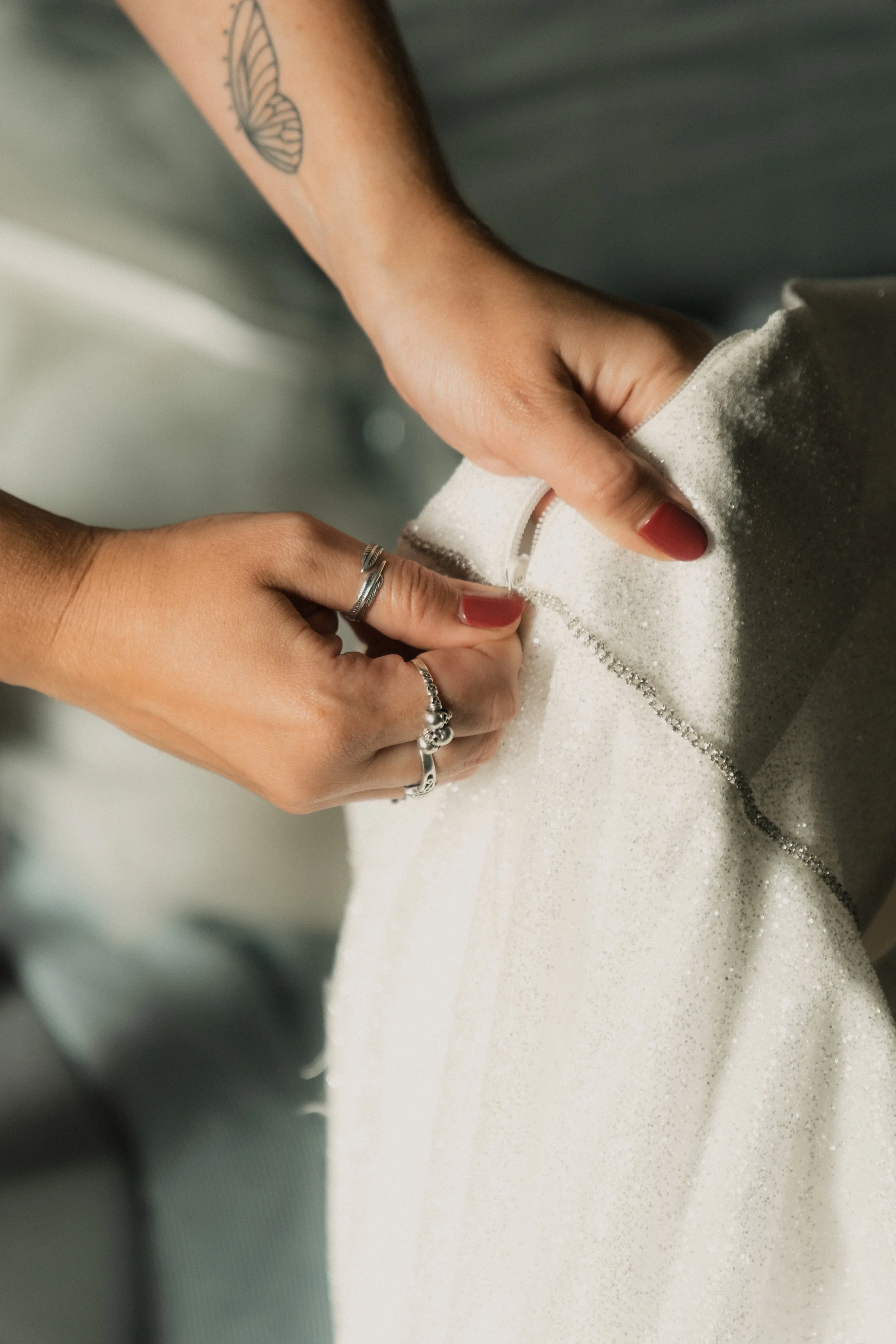 Femme en train de boutonner une robe de mariage blanche avec des doigts ornés de bijoux, on voit un tatouage papillon sur le bras.