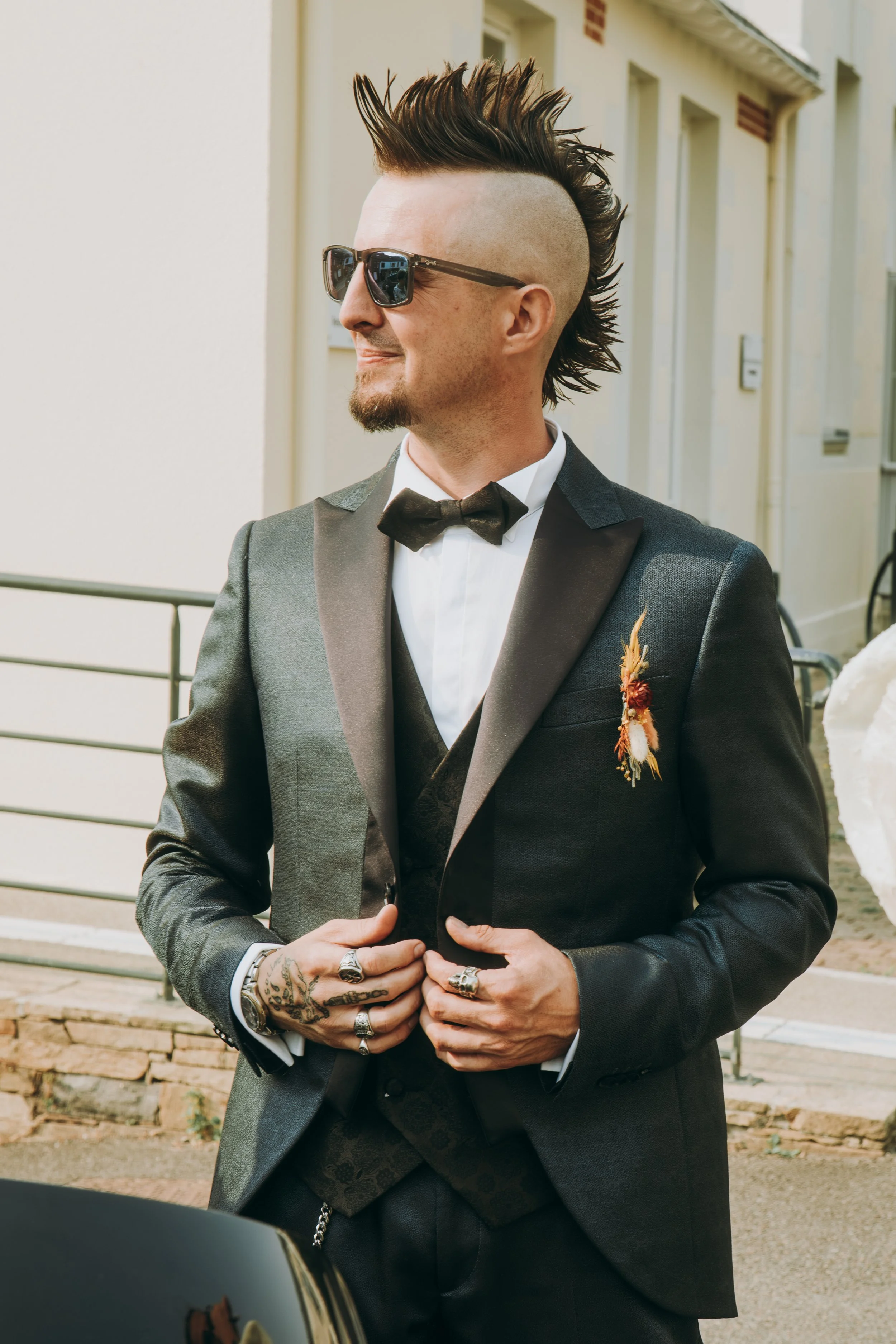 Homme avec une coiffure mohawk, lunettes de soleil, costume sombre, nœud papillon, et plusieurs bagues, posant à l'extérieur.