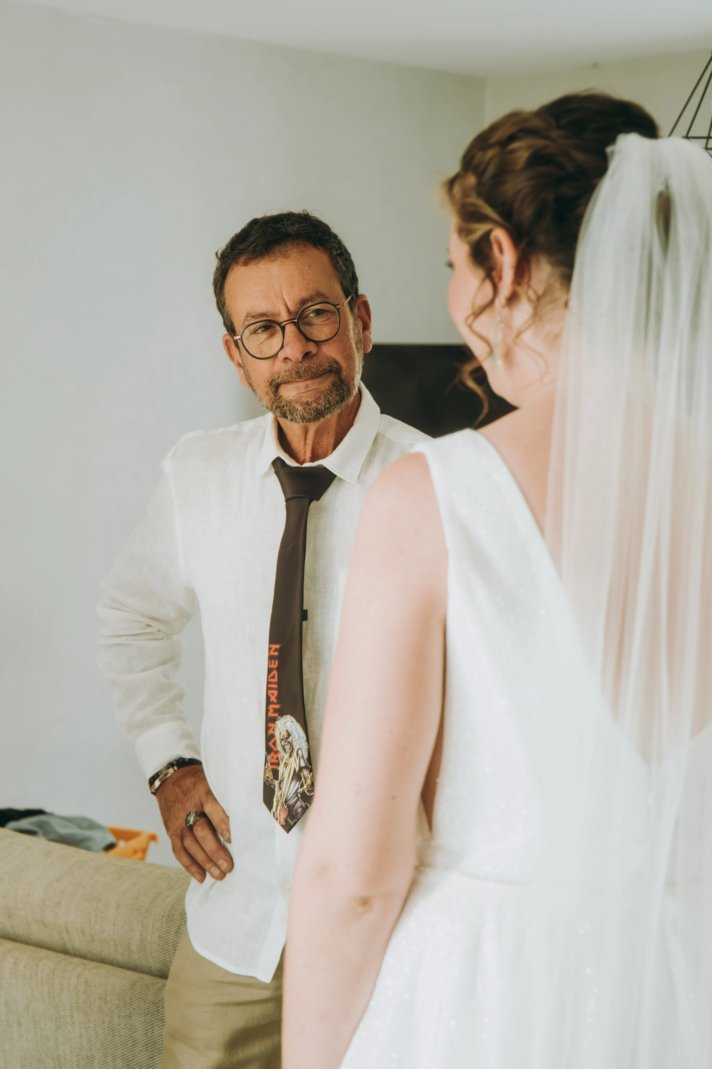 Un homme portant des lunettes, une chemise blanc et une cravate avec le logo de Iron Maiden, regarde la mariée lors d'un moment intime, dans une pièce intérieure.