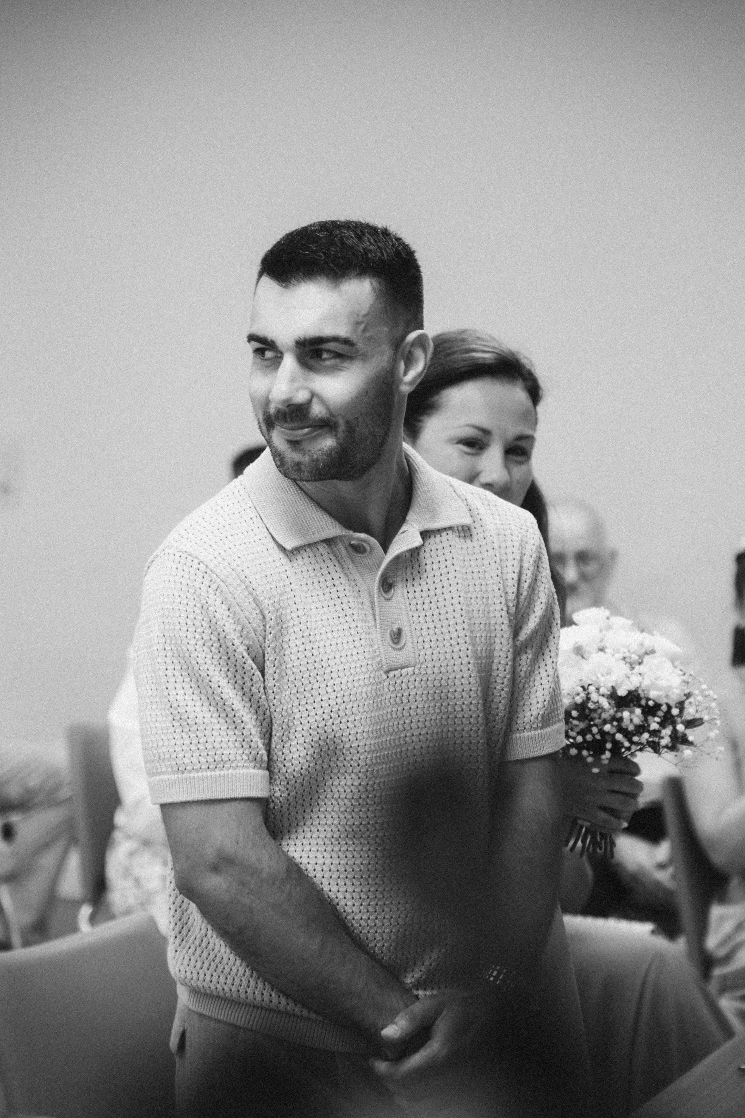Un homme en chemise à manches courtes, souriant, lors d'une cérémonie, avec une femme tenant un bouquet de fleurs en arrière-plan.