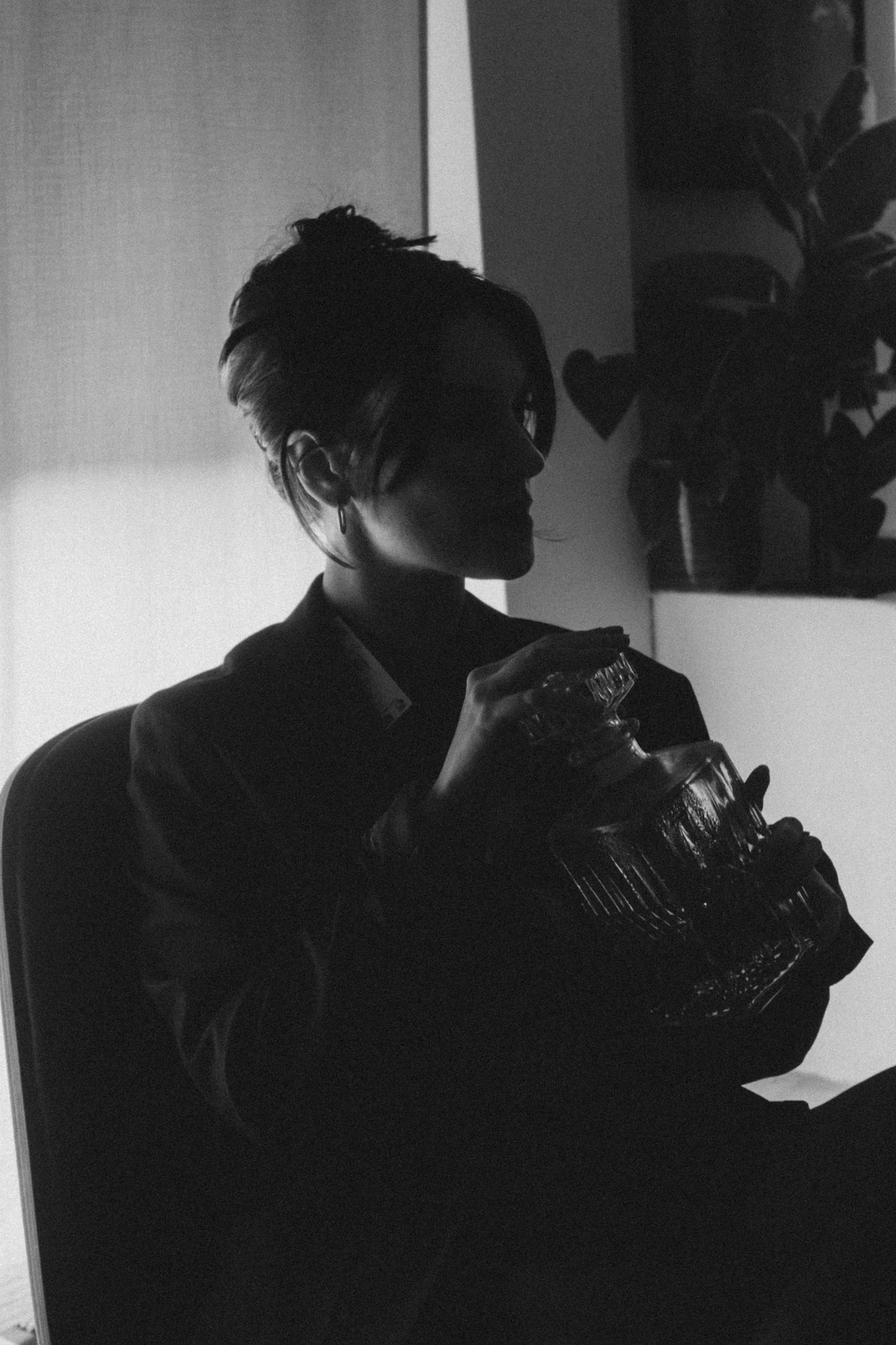 Silhouette d'une femme avec des cheveux short, tenant un bocal en verre, assise près d'une plante en pot, en intérieur.