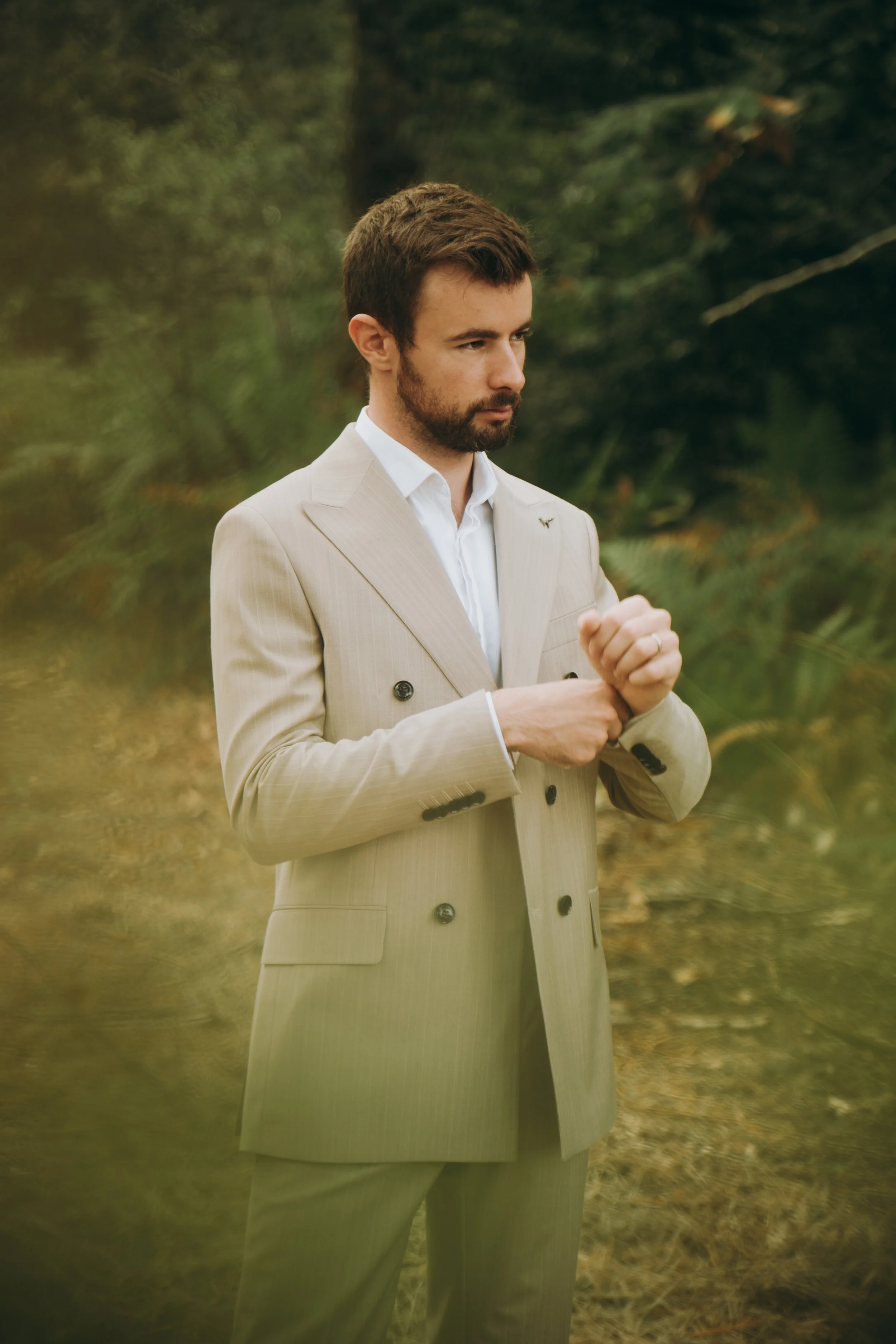Jeune homme en costume beige, assis dans un environnement naturel, probablement dans une forêt, regardant pensivement à droite.