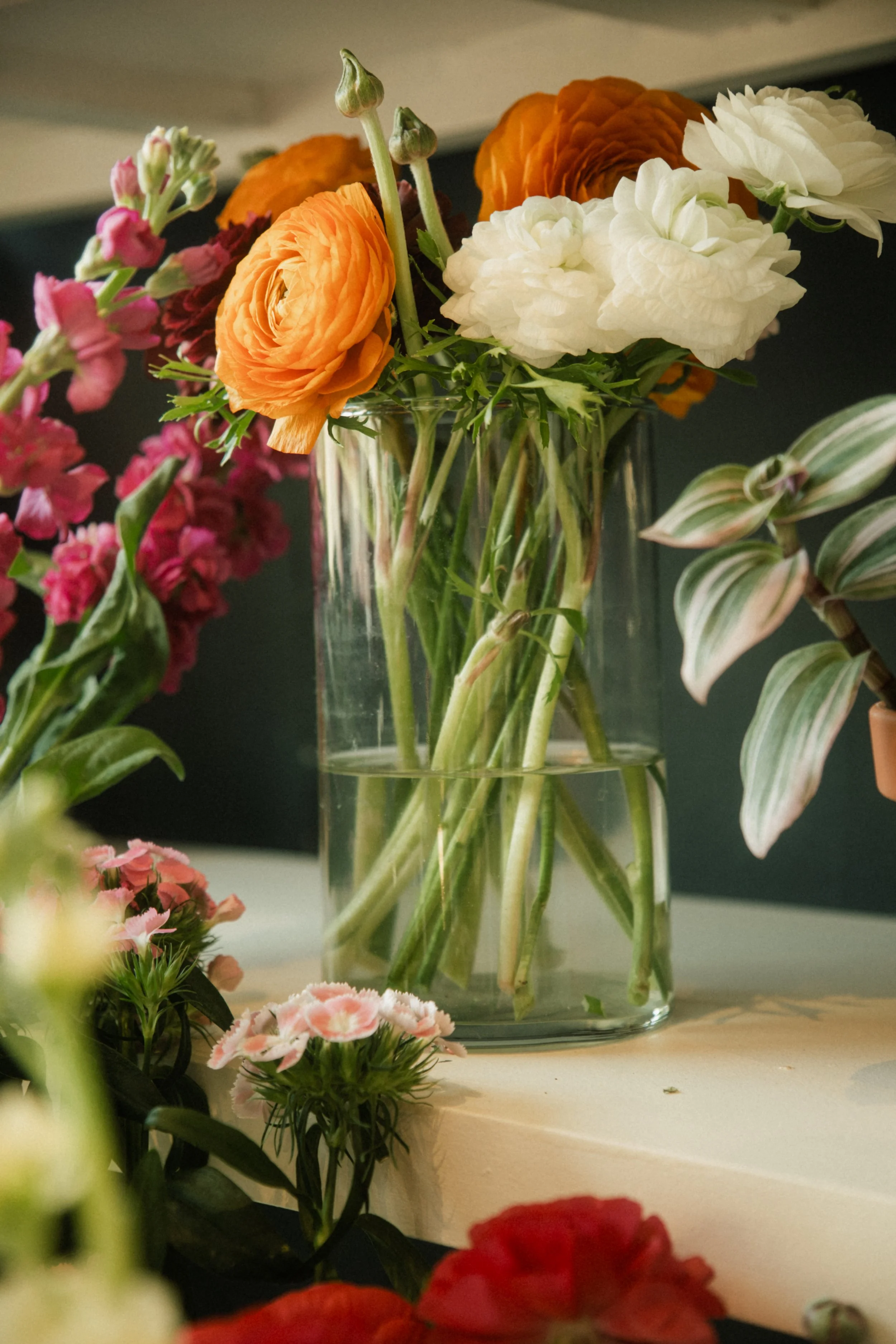 Un vase en verre contenant des fleurs de couleurs orange, blanc et rose, avec d'autres fleurs autour.