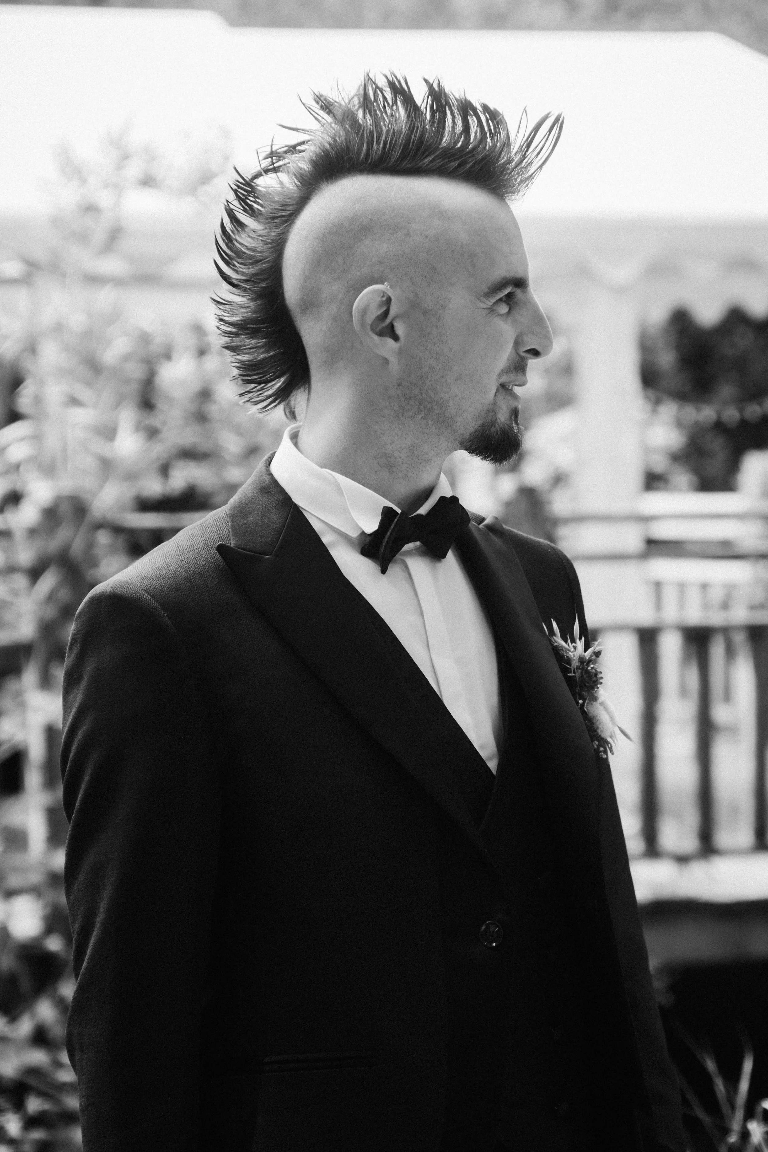 Un homme vêtu d'un costume de mariage avec une cravate papillon, en profil, contre un arrière-plan flou d'un balcon et de la végétation.