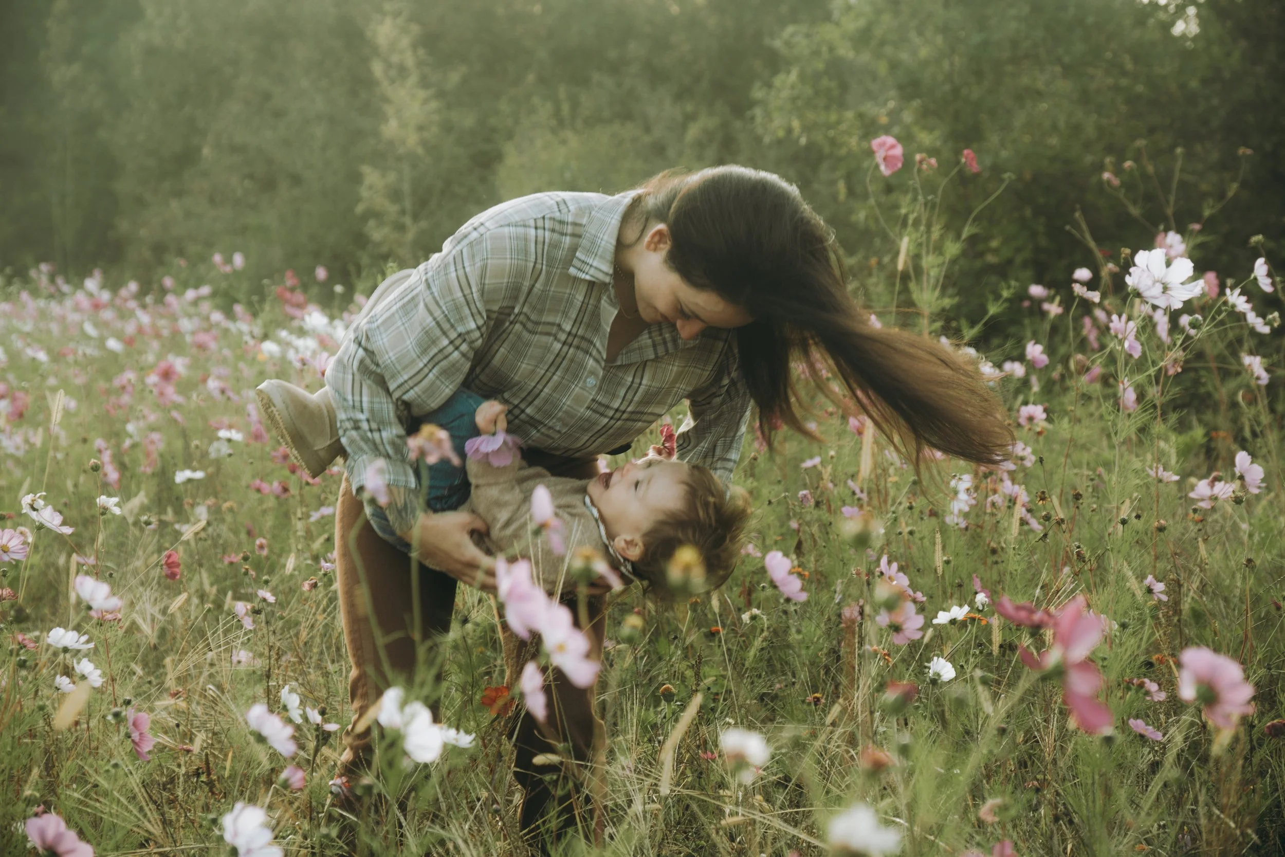 Une femme et un enfant jouent dans un champ de fleurs sauvages, la femme regarde l'enfant qui est allongé sur ses genoux, tous deux sourient dans un environnement naturel avec des fleurs roses et blanches.