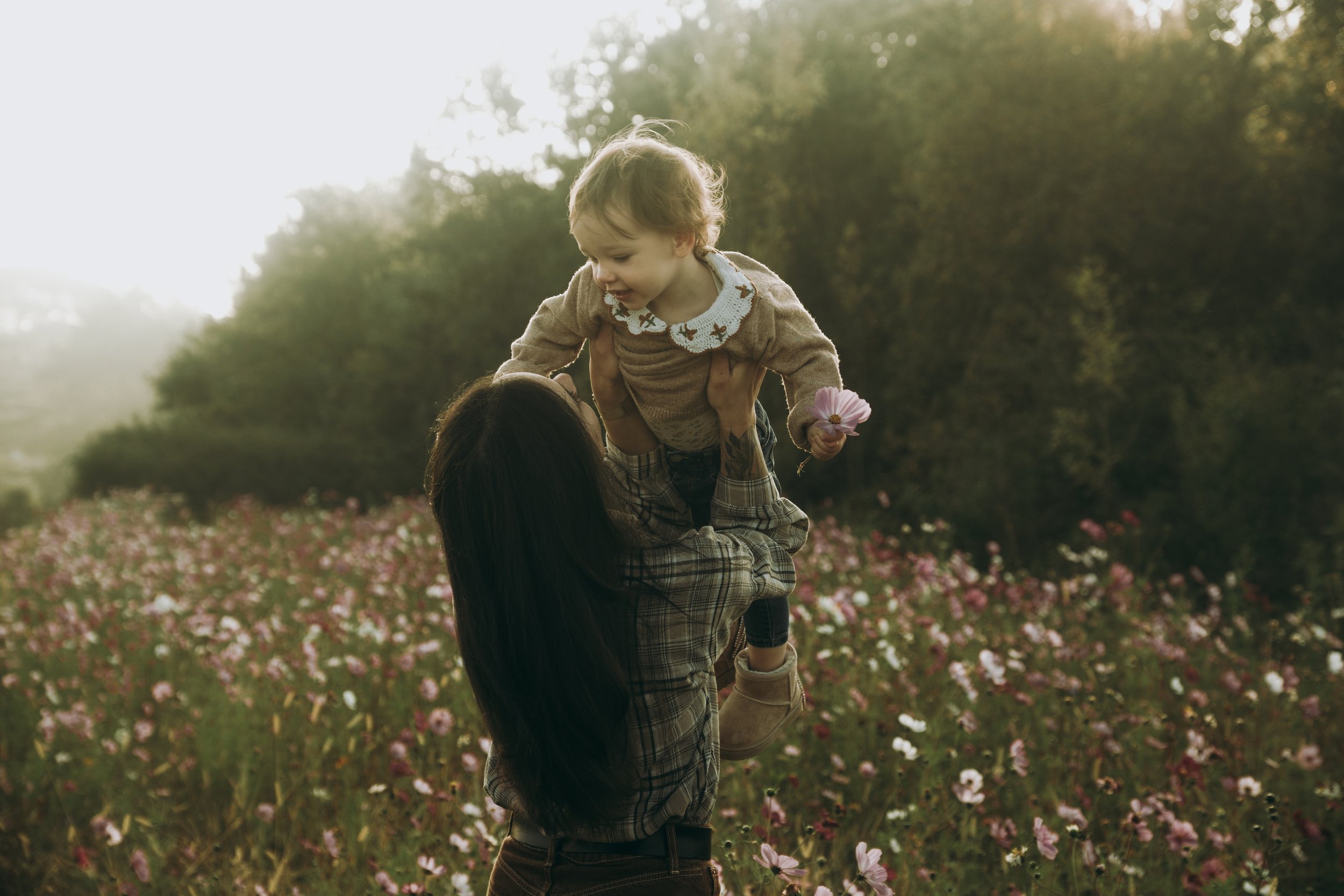 Une femme noire tenant un jeune garçon blond dans un champ de fleurs roses, avec des arbres en arrière-plan, lors d'un coucher de soleil.