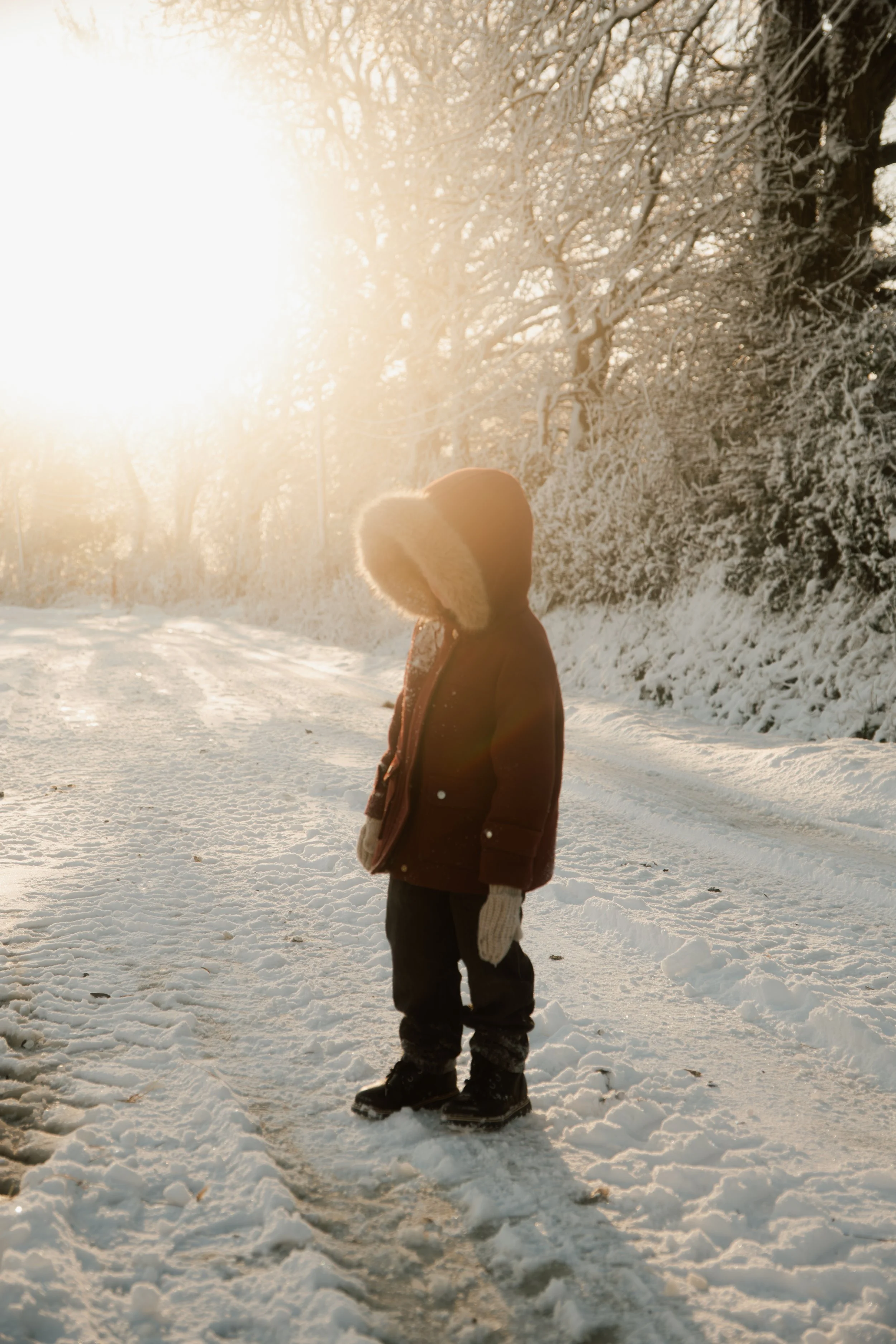 Enfant en manteau avec capuche en fourrure, gants et bottes sur un chemin enneigé en hiver, avec soleil brillant derrière les arbres.