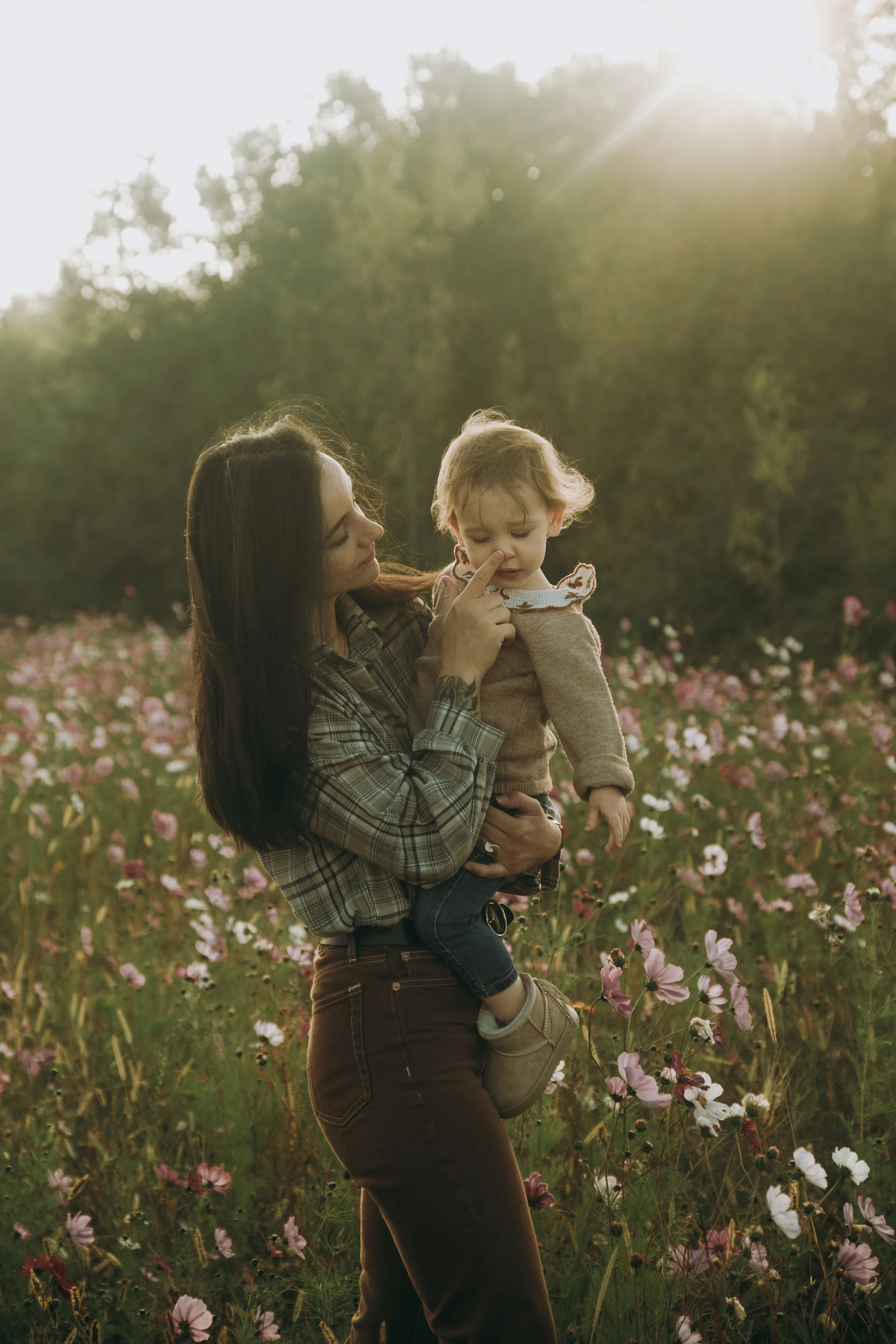 Une femme tient un enfant dans un champ de fleurs avec le soleil couchant en arrière-plan.