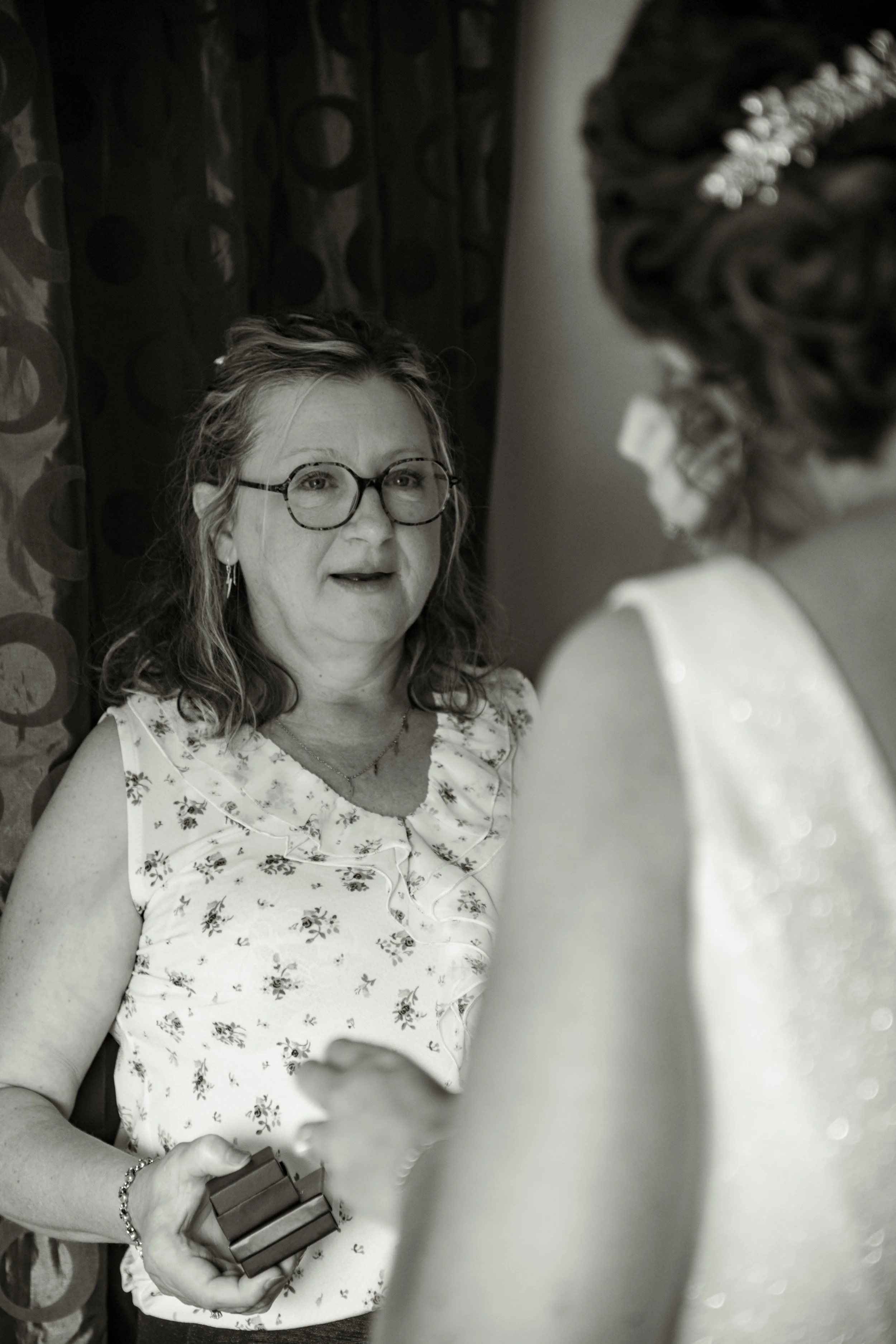 Une femme avec des lunettes et une robe à fleurs regarde une autre femme en robe de mariée lors d'une préparation de mariage.