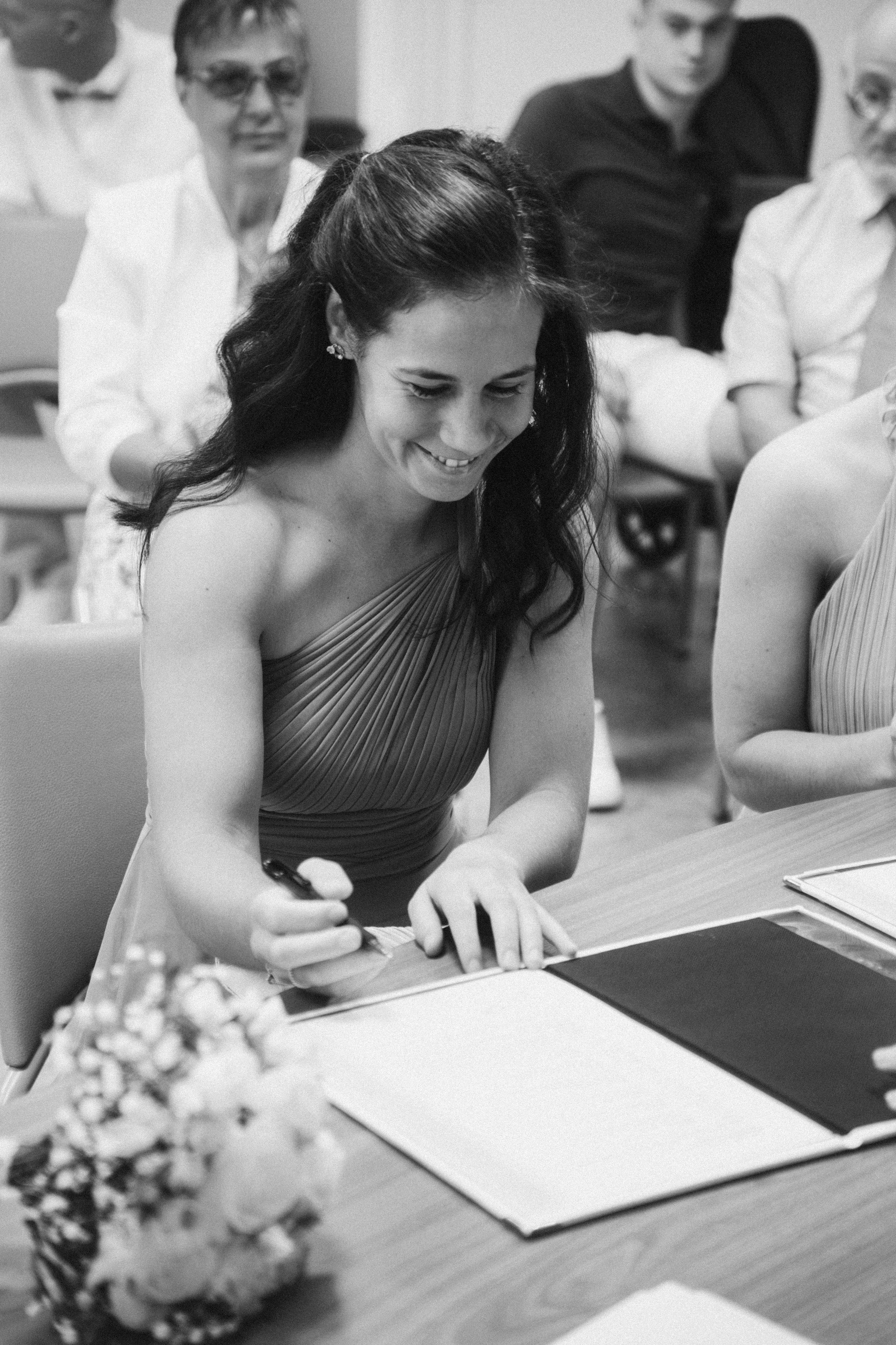 Une femme en robe plissée signature signant un document lors d'une cérémonie, entourée de plusieurs personnes qui l'observent, dans un environnement intérieur.