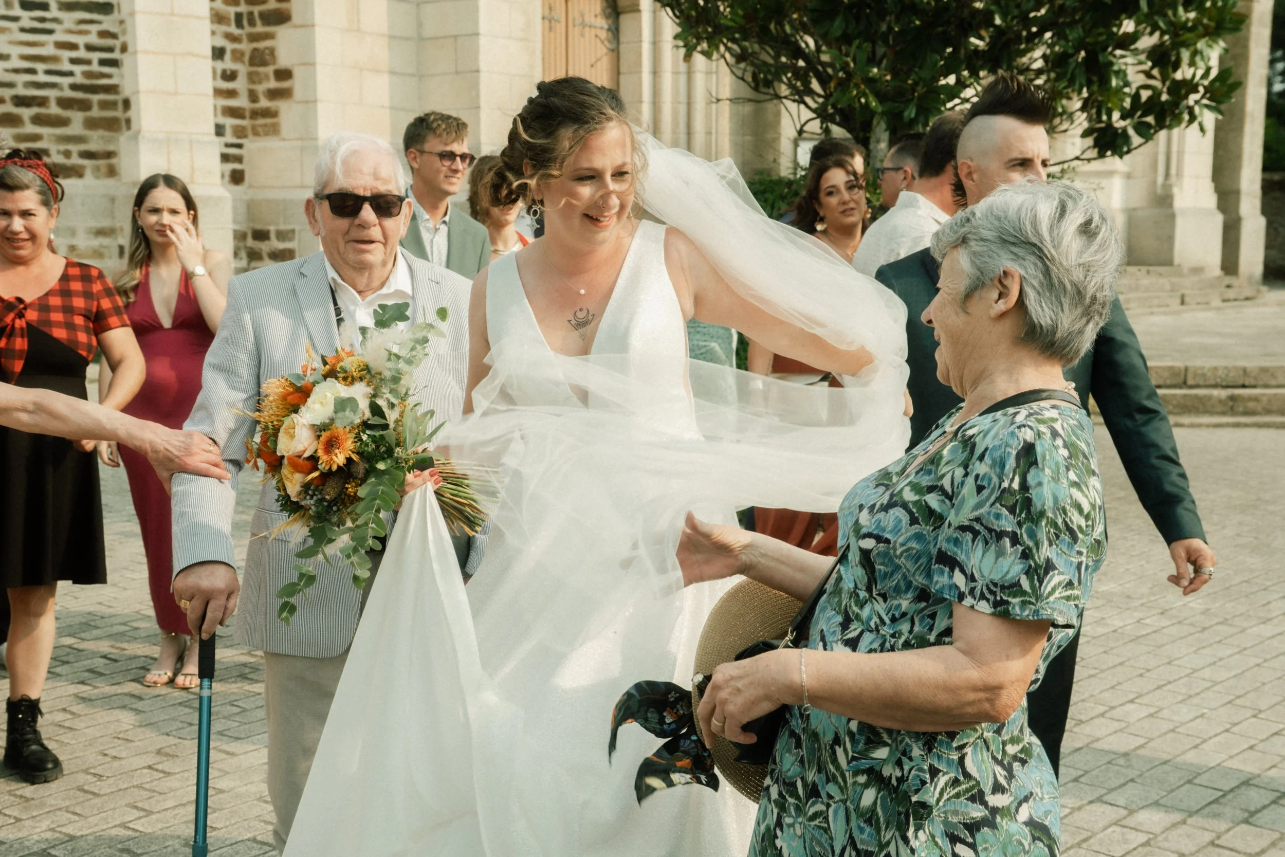 Une mariée souriante en robe blanche échangeant avec une femme âgée devant une église, entourée d'invités. La mariée tient un bouquet de fleurs colorées.