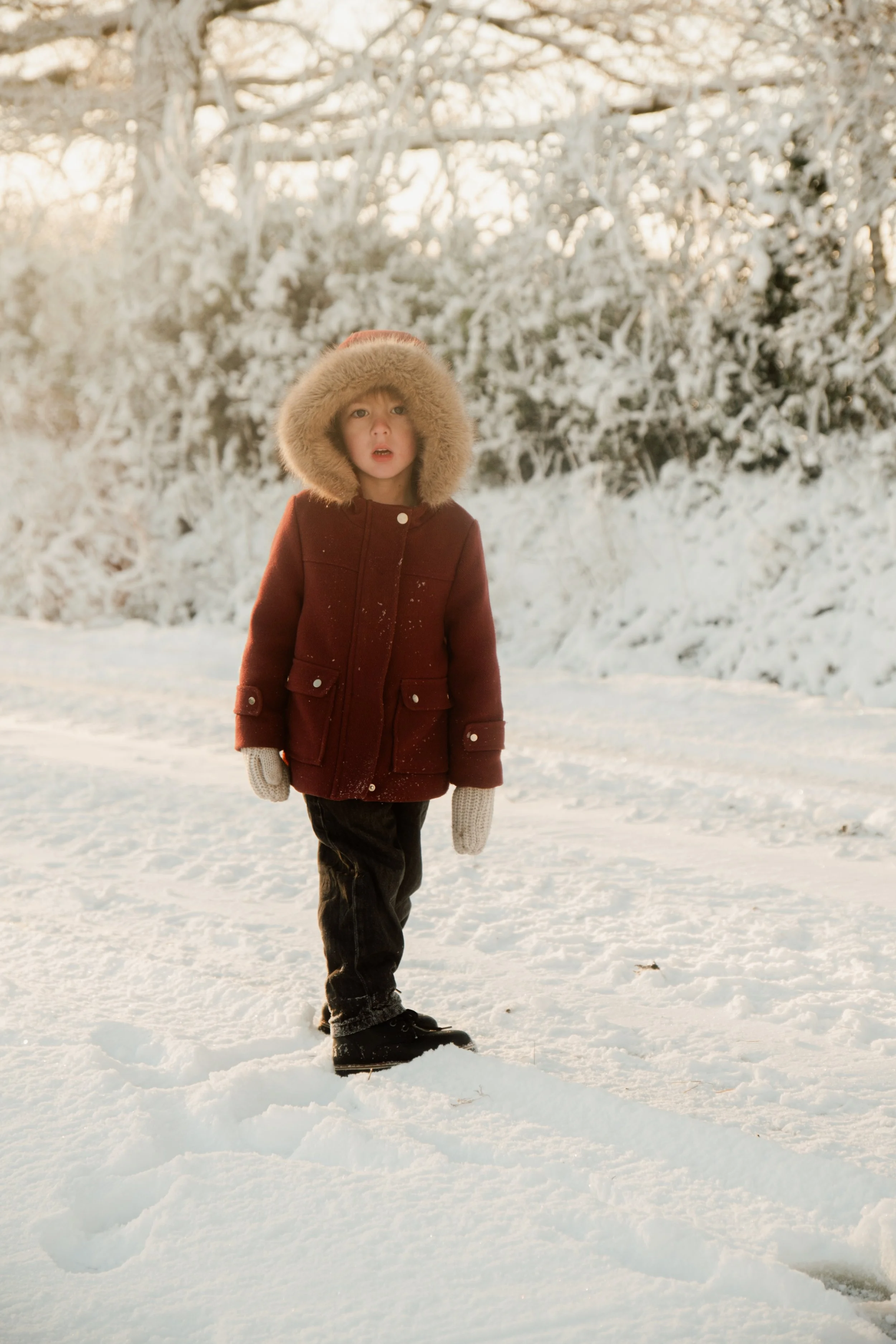 Un jeune garçon marche dans la neige, vêtu d'un manteau rouge avec une capuche en fourrure, des gants gris, et des chaussures noires, dans un paysage enneigé.