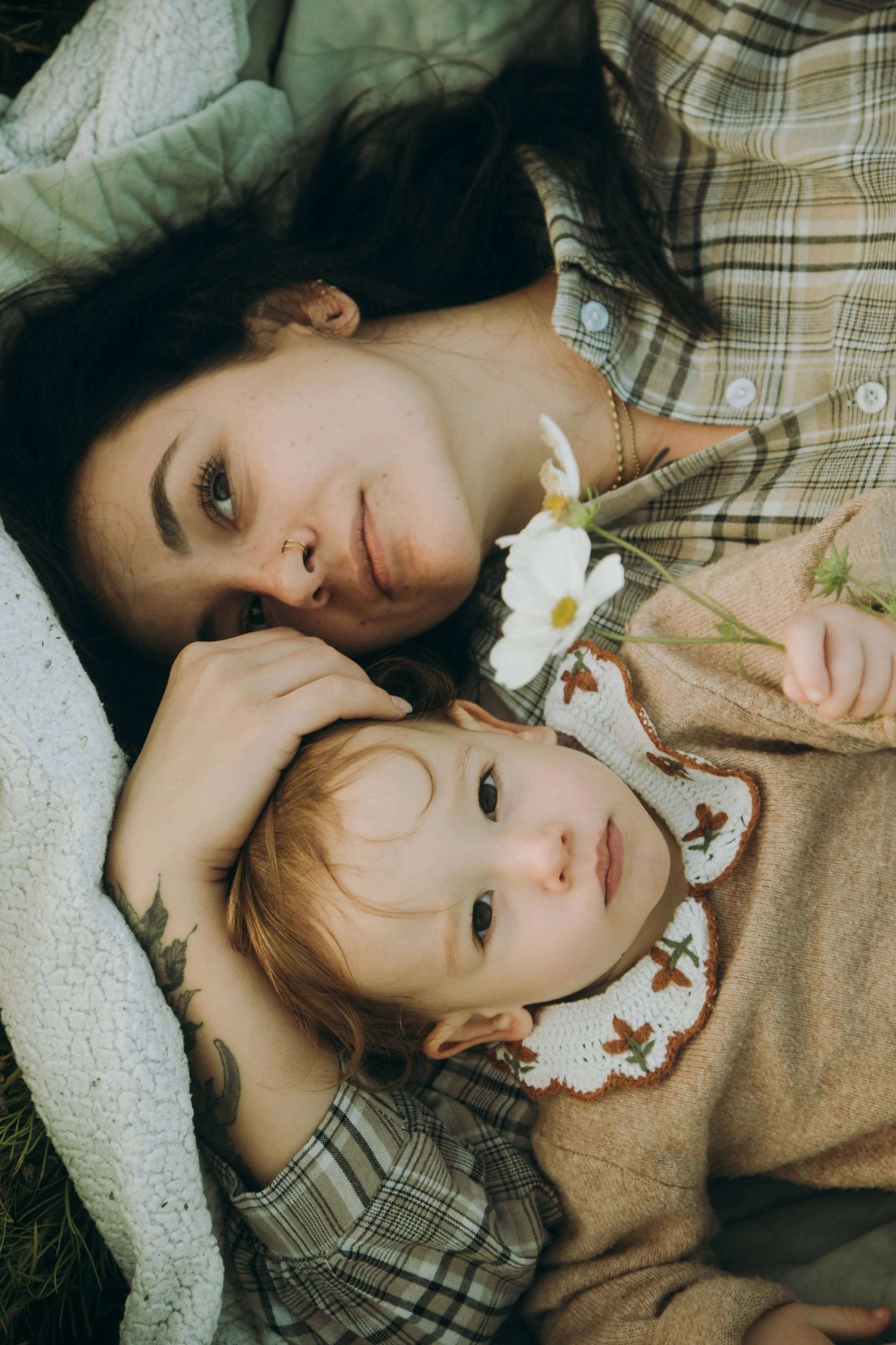 Une jeune femme avec des yeux bleus et une petite fille aux cheveux roux, allongées sur l'herbe, la femme tient une fleur blanche. Elles ont un look décontracté avec des vêtements à motifs.