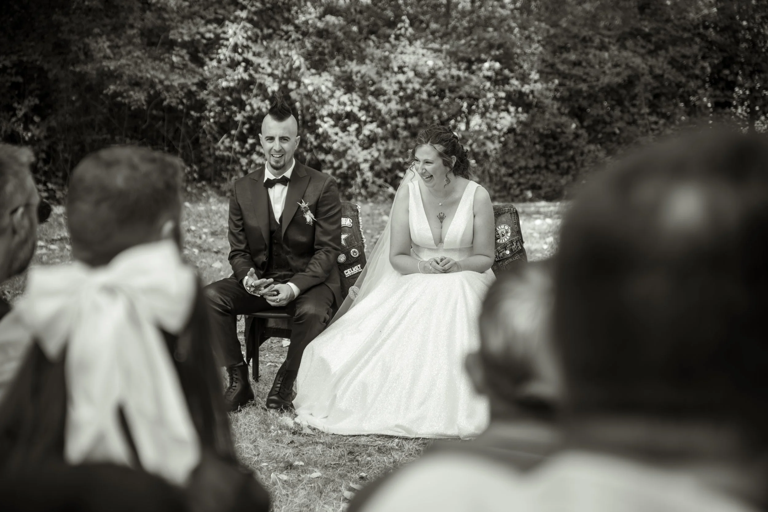 Un couple de mariage en plein air, la mariée en robe blanche et le marié en costume noir, assis et souriant, entourés d'invités. La scène est dans un environnement naturel avec des arbres en arrière-plan.