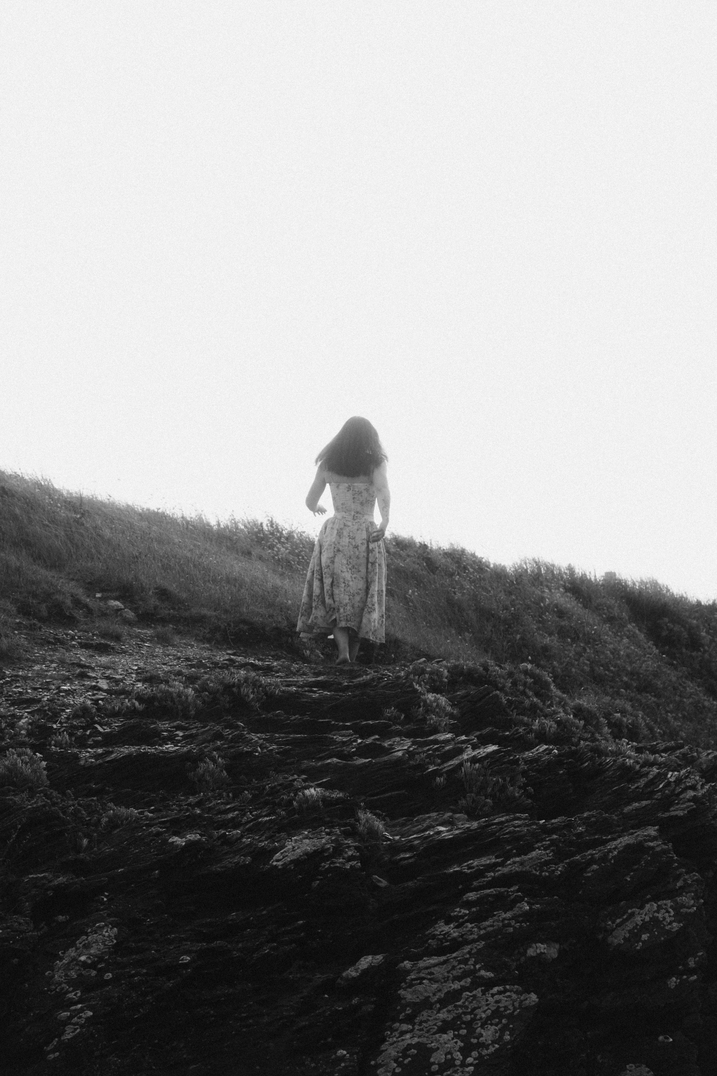 Une femme en robe longue marche sur un sentier en pente sur une colline, vue en noir et blanc.