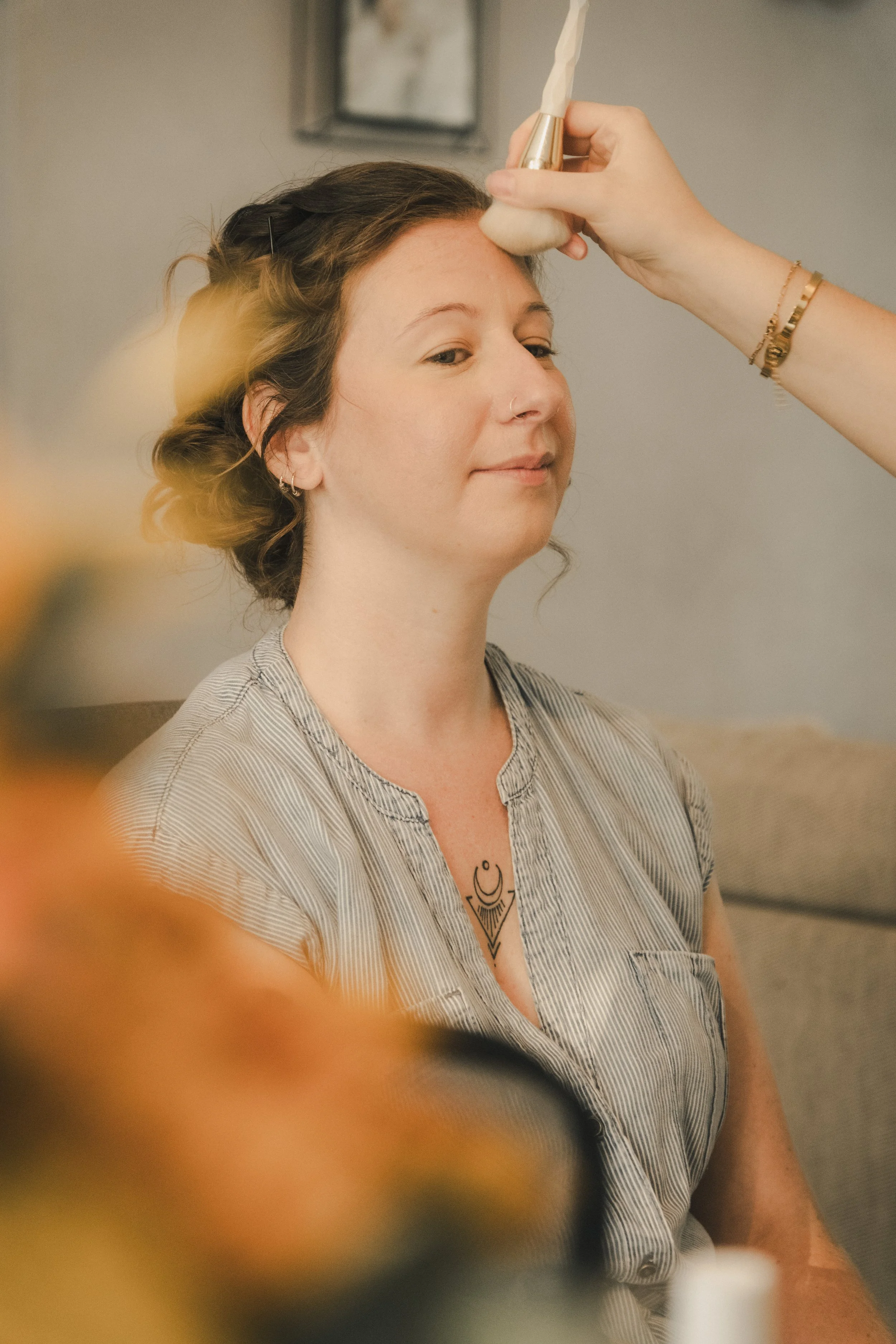 Une femme est en train de se faire maquiller le front avec un pinceau par une autre personne, dans un cadre intérieur.