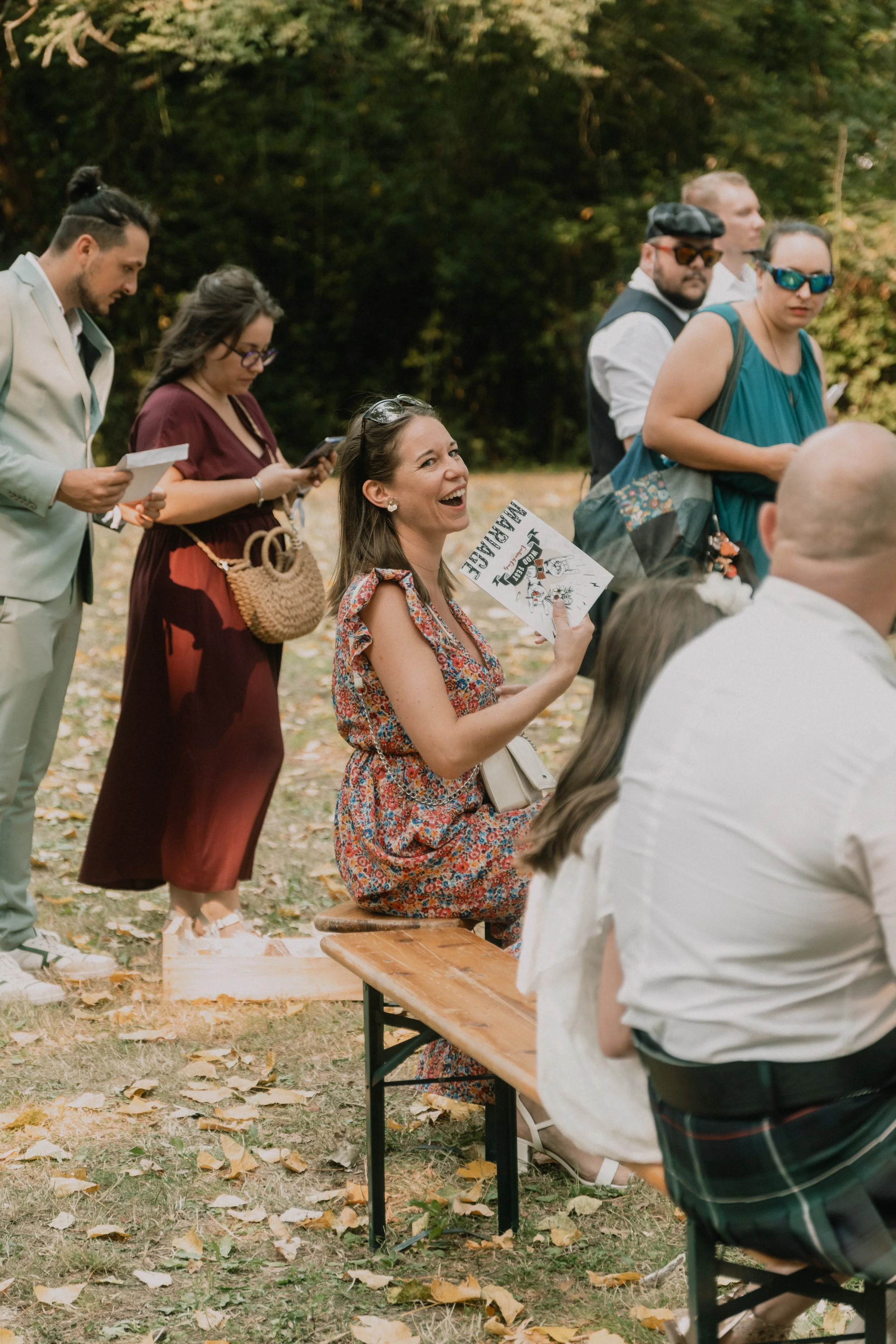 Un groupe de personnes assises et debout lors d'un événement en plein air, sous les arbres, avec une femme souriante au centre tenant une affiche.
