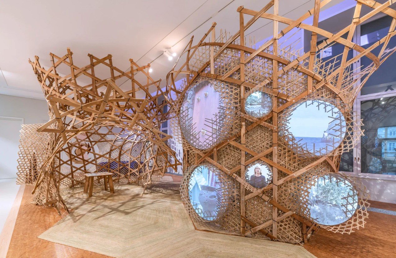 A contemporary art installation made of interwoven wooden latticework, featuring large circular mirrors and multiple viewing windows, inside a room with large windows and a wooden floor.