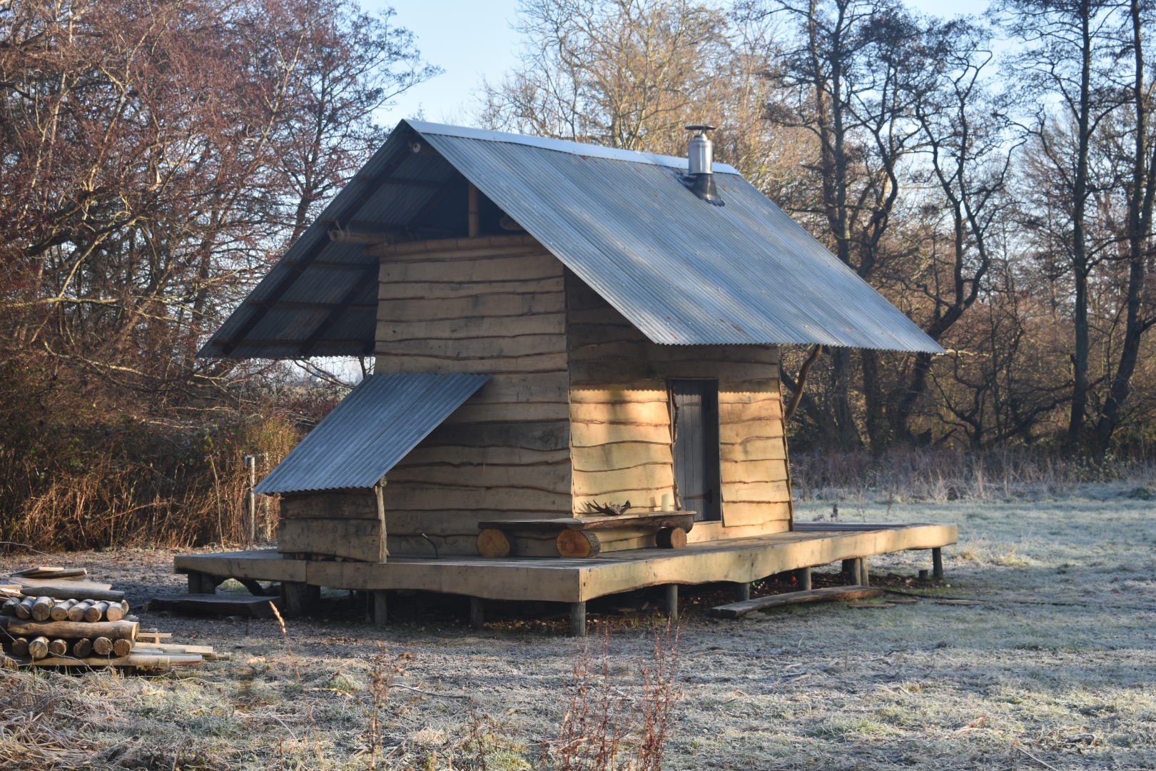 A small wooden house with a corrugated metal roof, on a raised platform in a frosty field with trees in the background.