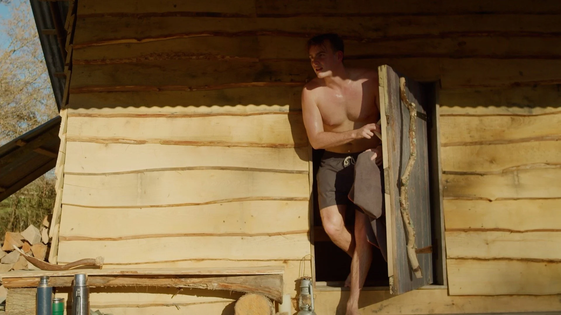 A shirtless man in black shorts standing at a small wooden cabin window, leaning out and looking outside.