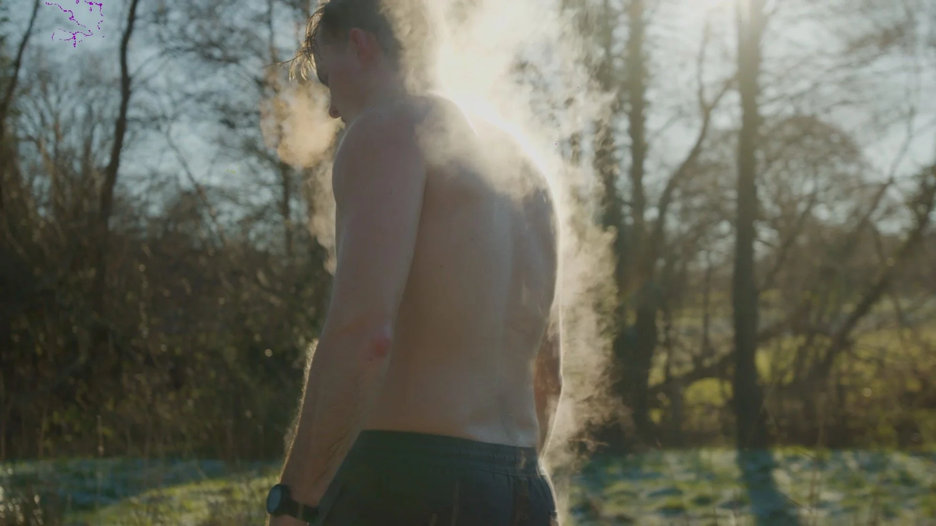 A shirtless man with a smartwatch on his left wrist standing outdoors among trees, with steam or breath visible in cold weather, backlit by sunlight.