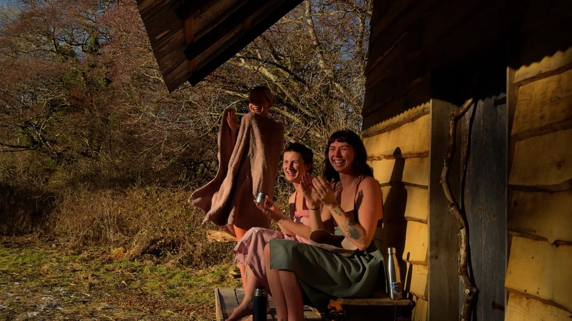Three women sitting on a porch of a rustic wooden cabin, laughing and enjoying an outdoor moment. One woman is standing and waving, with a towel or blanket over her shoulders. Two women are seated, clapping and smiling. The scene is sunny with trees in the background.