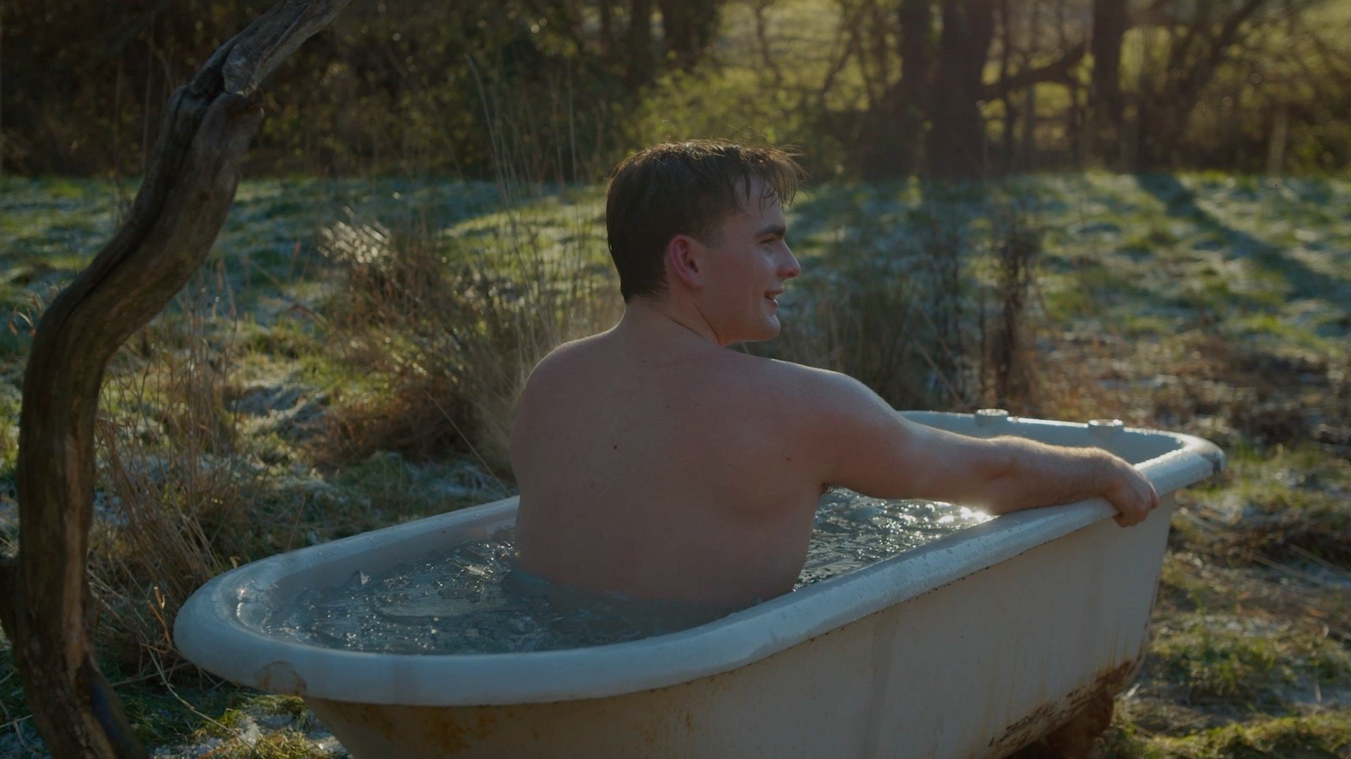 A young man with short brown hair enjoying a bath in an outdoor bathtub in a natural setting with trees and grass, water splashing from a wooden water spout