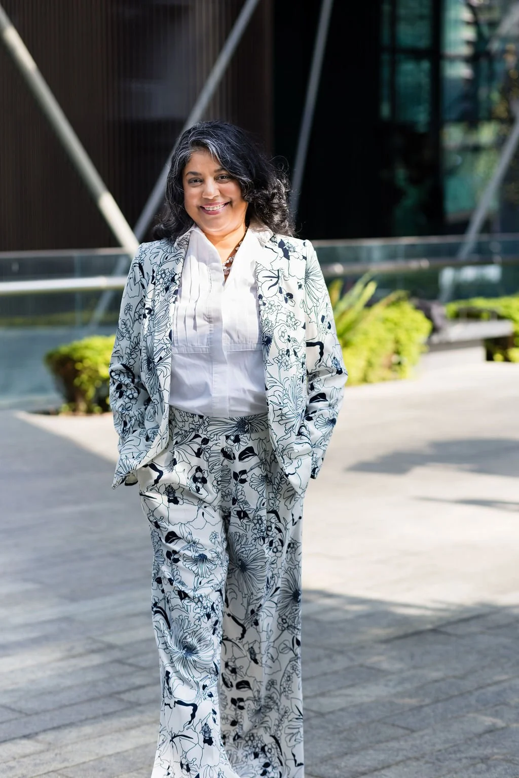 Woman in a floral suit and white blouse walking outdoors in bright sunlight, with modern building and greenery in the background.
