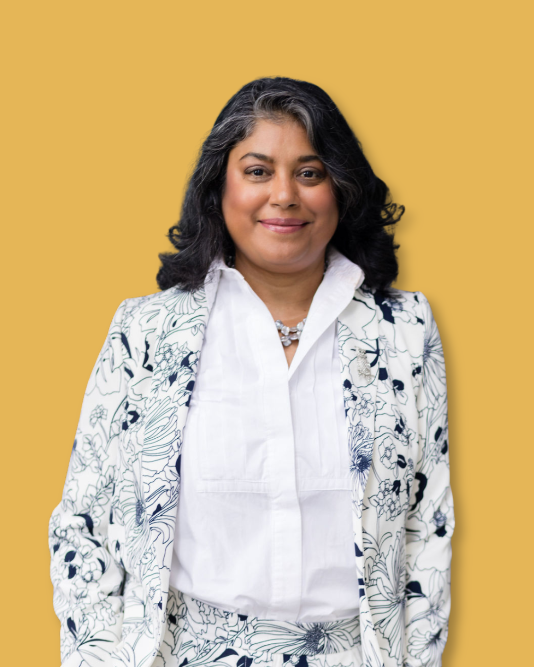 A woman with black hair and a white floral blazer. She is smiling and wearing a white blouse and a pearl necklace, standing against a mustard yellow background.