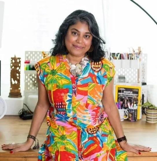 Woman with dark hair in colorful tropical dress standing in a bright room with plants and office supplies.
