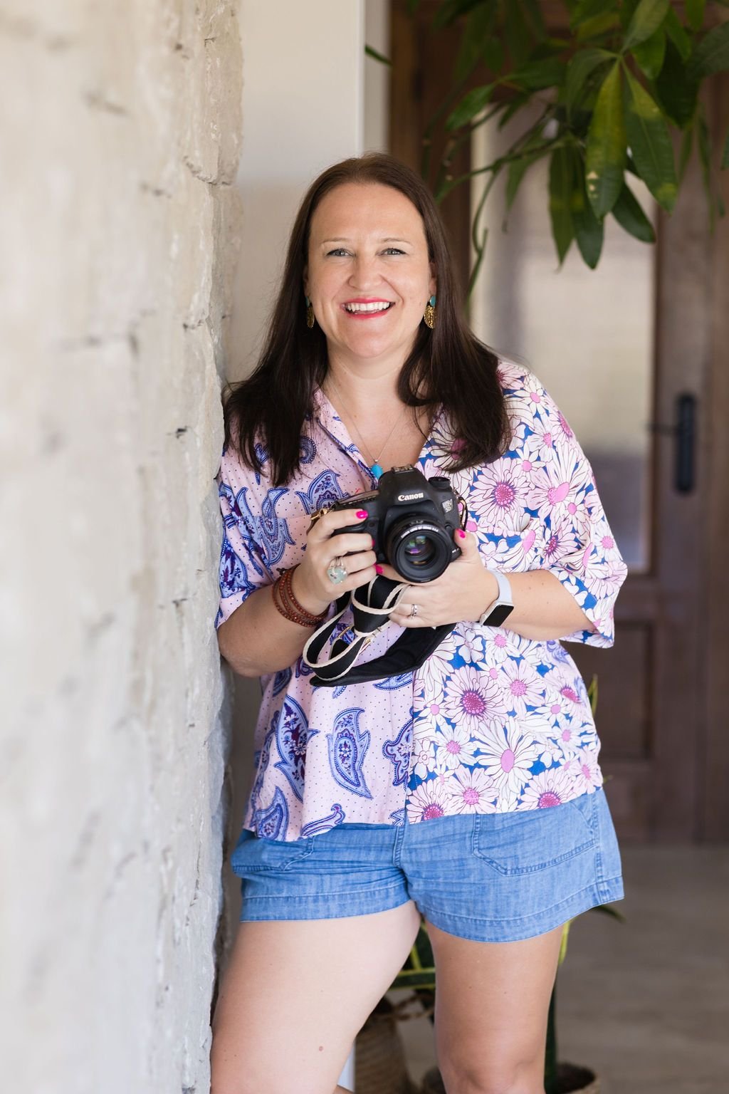 A woman with long dark hair smiling, holding a Canon camera, standing next to a stone wall with a plant in the background, dressed in a floral shirt and denim shorts.