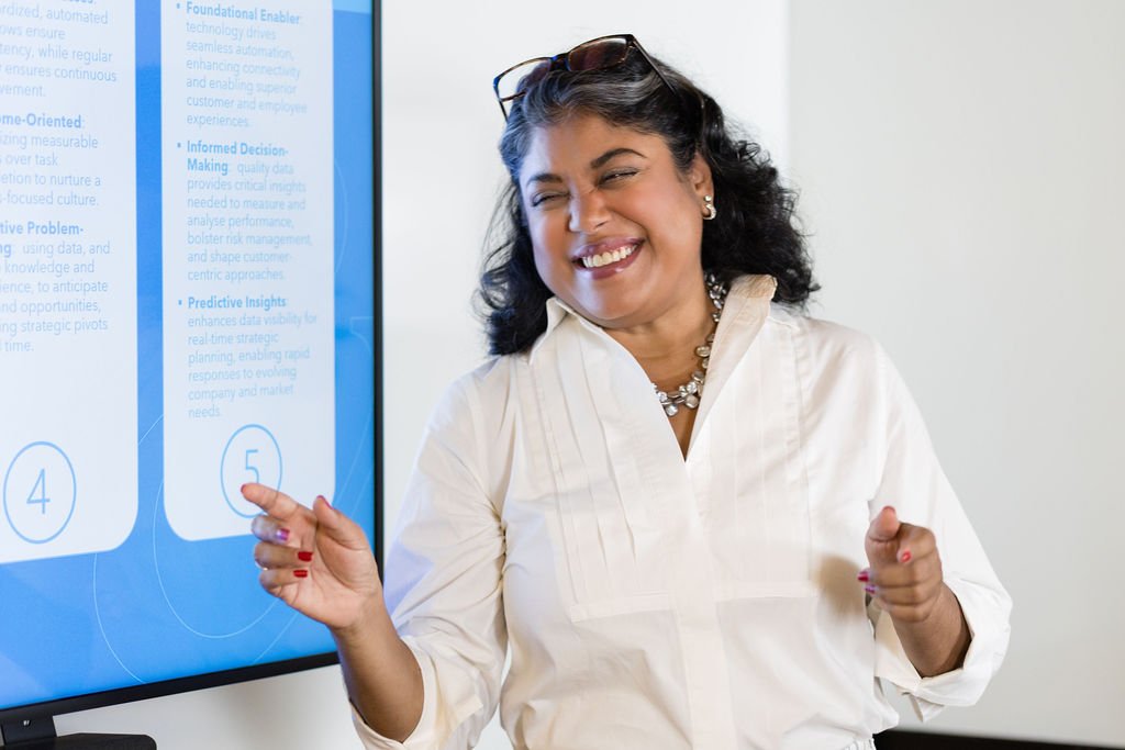 A woman smiling and pointing at a large screen with presentation slides, in business attire, wearing earrings and a necklace.