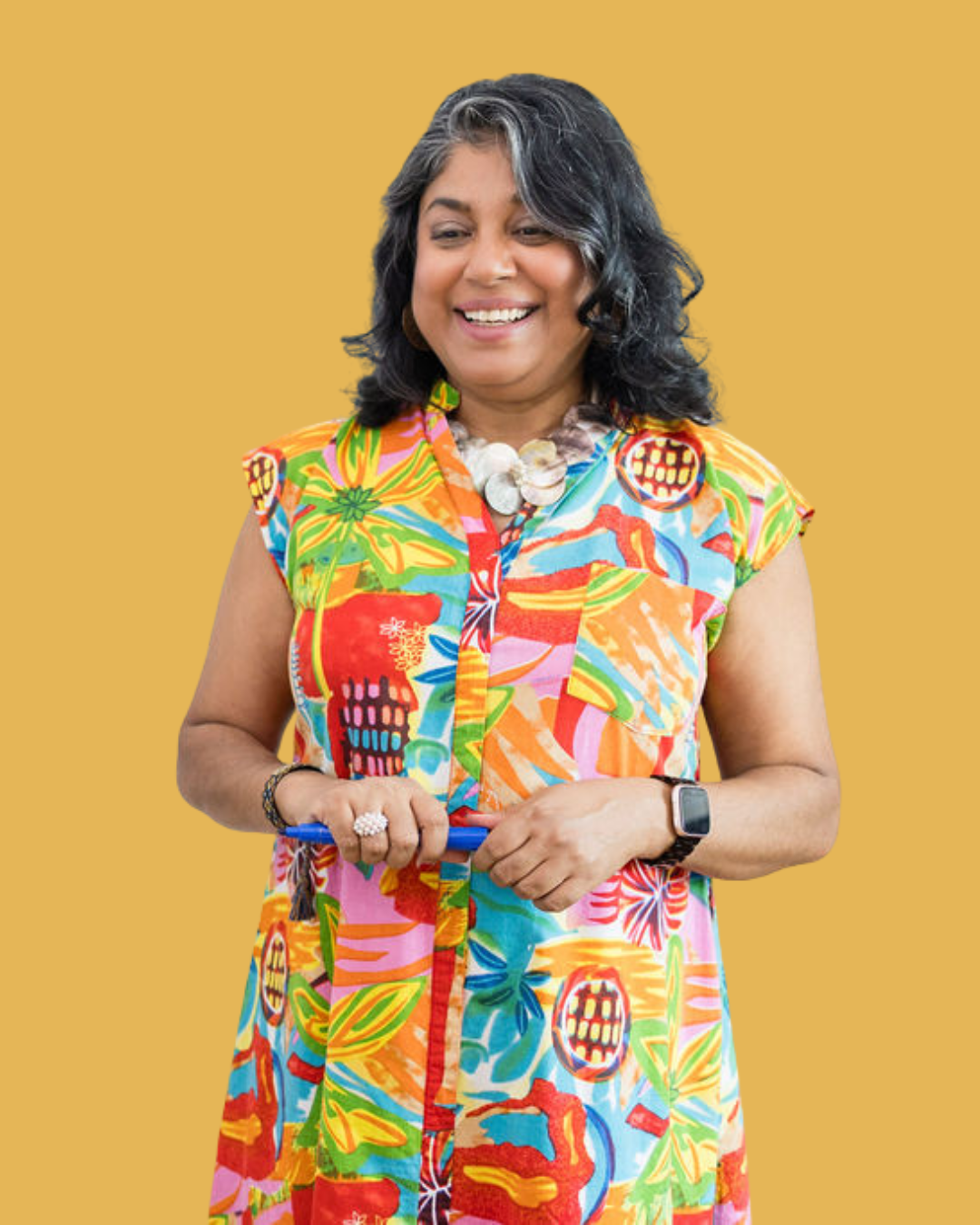 A woman smiling, wearing a colorful, tropical-patterned dress, a shell necklace, and a smartwatch, standing against a yellow background.