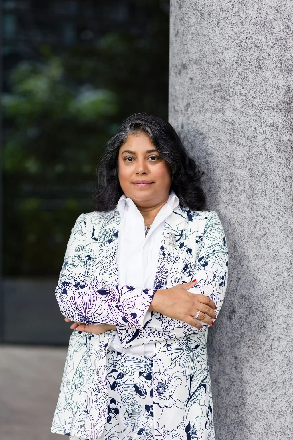 A woman with dark, curly hair and medium skin tone wearing a white floral suit with a collared shirt, standing with arms crossed against a gray textured wall with blurred greenery in the background.