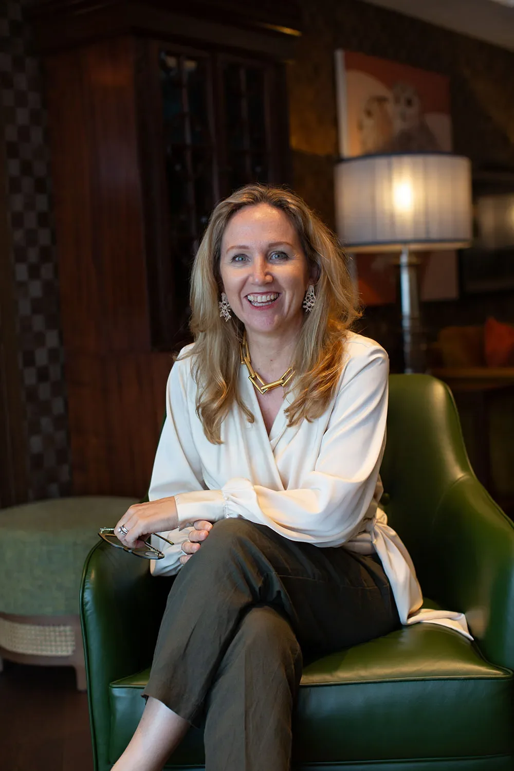 A smiling woman with blonde hair, wearing a white blouse, dark pants, and jewelry, sits on a green armchair in a warmly lit living room with wooden furniture and artwork in the background.